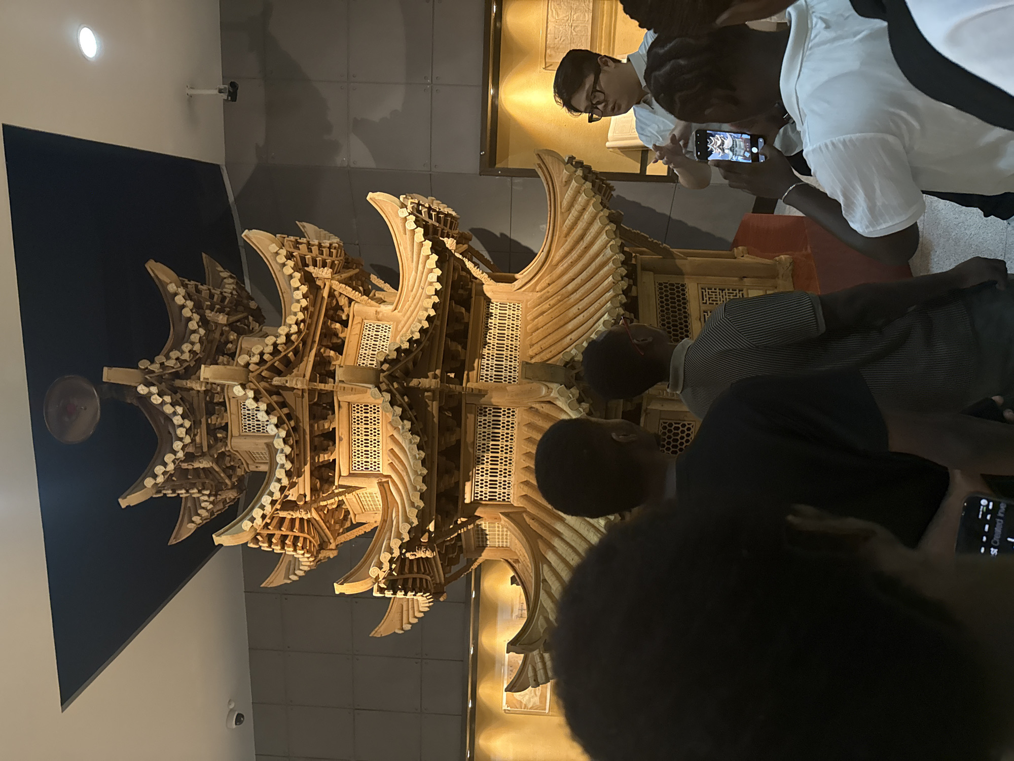 In a museum setting, likely in Yuzhong District, China, a group of young people are observing a detailed, multi-tiered wooden model of a traditional Chinese pagoda. The pagoda is intricately carved with curved roofs and latticed windows, illuminated by warm lighting that casts shadows on the surrounding walls. Several individuals are actively engaged with the exhibit. One person, wearing glasses and a light-colored shirt, leans in to examine the structure. Another, holding a smartphone, is taking a picture of the pagoda. The people are dressed casually, suggesting a daytime visit. The atmosphere appears relaxed and engaged, typical of a museum visit. The background shows a muted, possibly grey, wall with some framed artwork, and a security camera is visible on the wall. The lighting suggests it is likely indoors, possibly evening or a dimly lit daytime.