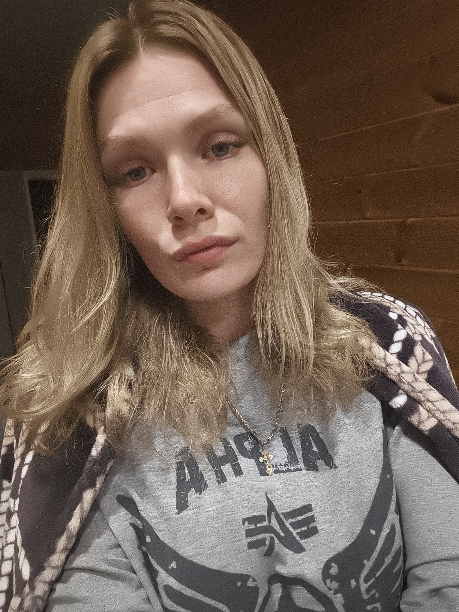 A woman with long blonde hair looks directly at the camera. She wears a gray long-sleeved shirt with a black Alpha Industries logo. A brown and white patterned shawl drapes over her shoulders. A gold necklace with a cross pendant hangs around her neck. The background is a wood-paneled wall and a partially visible doorway. The location is likely indoors, but not definitively identifiable as Русский Вожой, Russia.