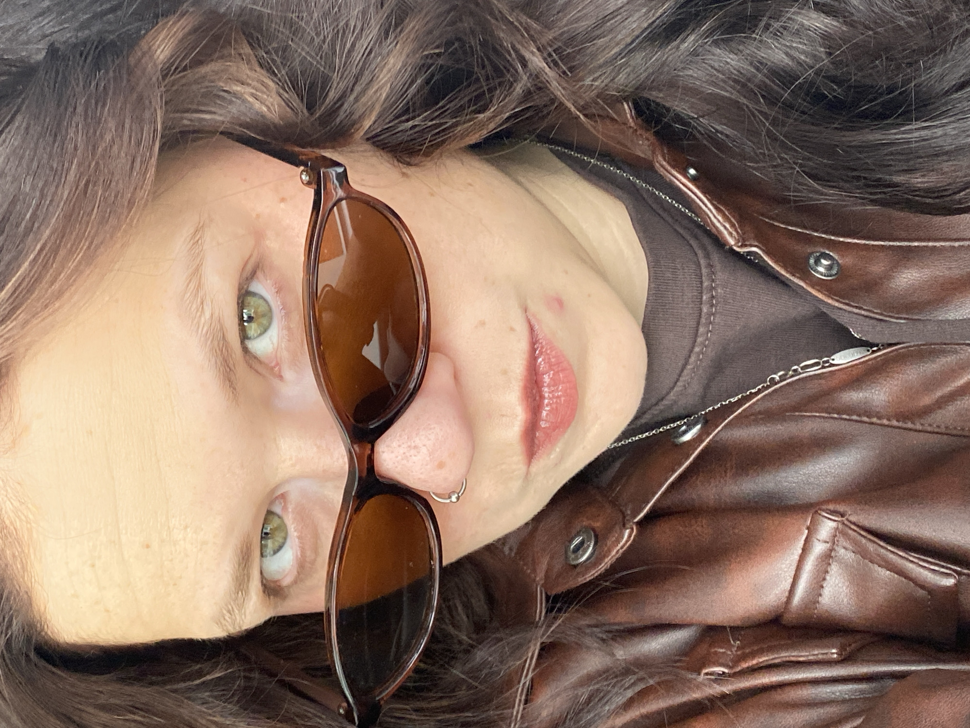 A close-up portrait shows a person with medium-brown, wavy hair and distinct light green eyes, oriented sideways in the frame. Their head is tilted, and they appear to be looking towards the viewer. The person has fair skin, a neutral expression with slightly parted lips, and a glossy pinkish-brown lip color.
They are wearing dark brown, tortoiseshell-patterned sunglasses that rest on their nose, partially covering their eyes. A small silver ring nose piercing is visible on their left nostril, and a small blemish is present on the right cheek. The individual is dressed in a dark brown top, possibly a t-shirt, visible beneath a reddish-brown leather or faux-leather jacket. The jacket features snap buttons and a zipper detail around the collar and chest area. A thin silver chain necklace is worn, and a small, unreadable silver tag is attached to it. The person's hair is voluminous and frames their face. Natural light illuminates the scene, creating reflections in the sunglasses lenses and the person's eyes. No specific activity beyond posing is depicted. The image does not contain any discernible text, apart from the unreadable tag on the necklace.