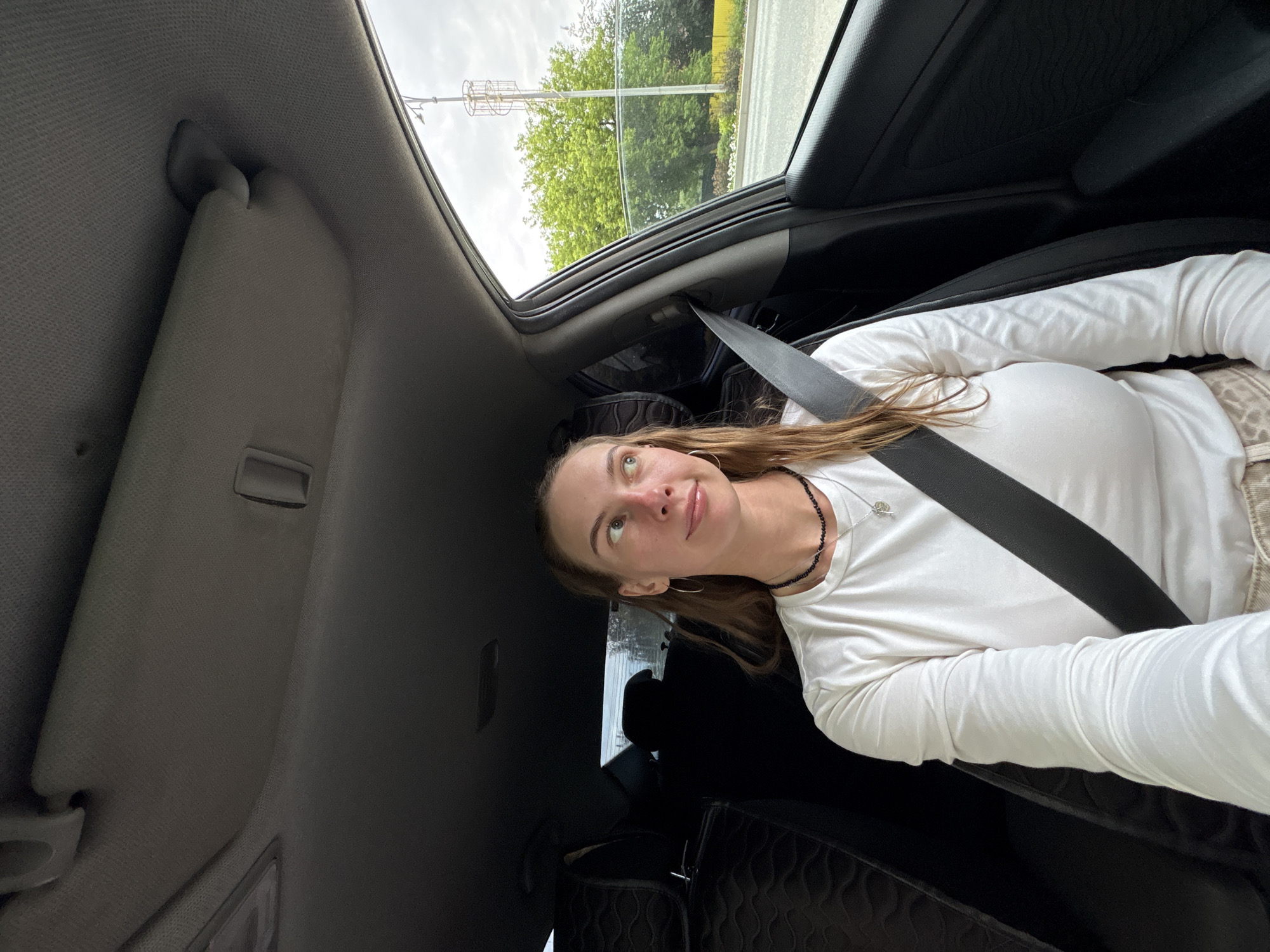 A young woman with long, light brown hair and fair skin is captured in a selfie taken from inside a car. She is wearing a white long-sleeved shirt and a black beaded necklace. A seatbelt is fastened across her chest. Her eyes are looking upwards and to the side, with a slight smile on her face, conveying a sense of contemplation or mild amusement.
The scene is the interior of a car, with the sunroof open, revealing a bright, overcast sky. Trees and a utility pole are visible through the open sunroof, suggesting an outdoor setting. The car's interior, including the gray headliner and black trim, is visible. A quilted black bag is on the seat behind the woman.
The image appears to be taken during the daytime, under cloudy weather conditions. The location is indicated to be Perm, Russia, although there are no explicit text cues or landmarks within the image to confirm this. The overall mood is relaxed and casual, suggesting the woman is a passenger enjoying a car ride.