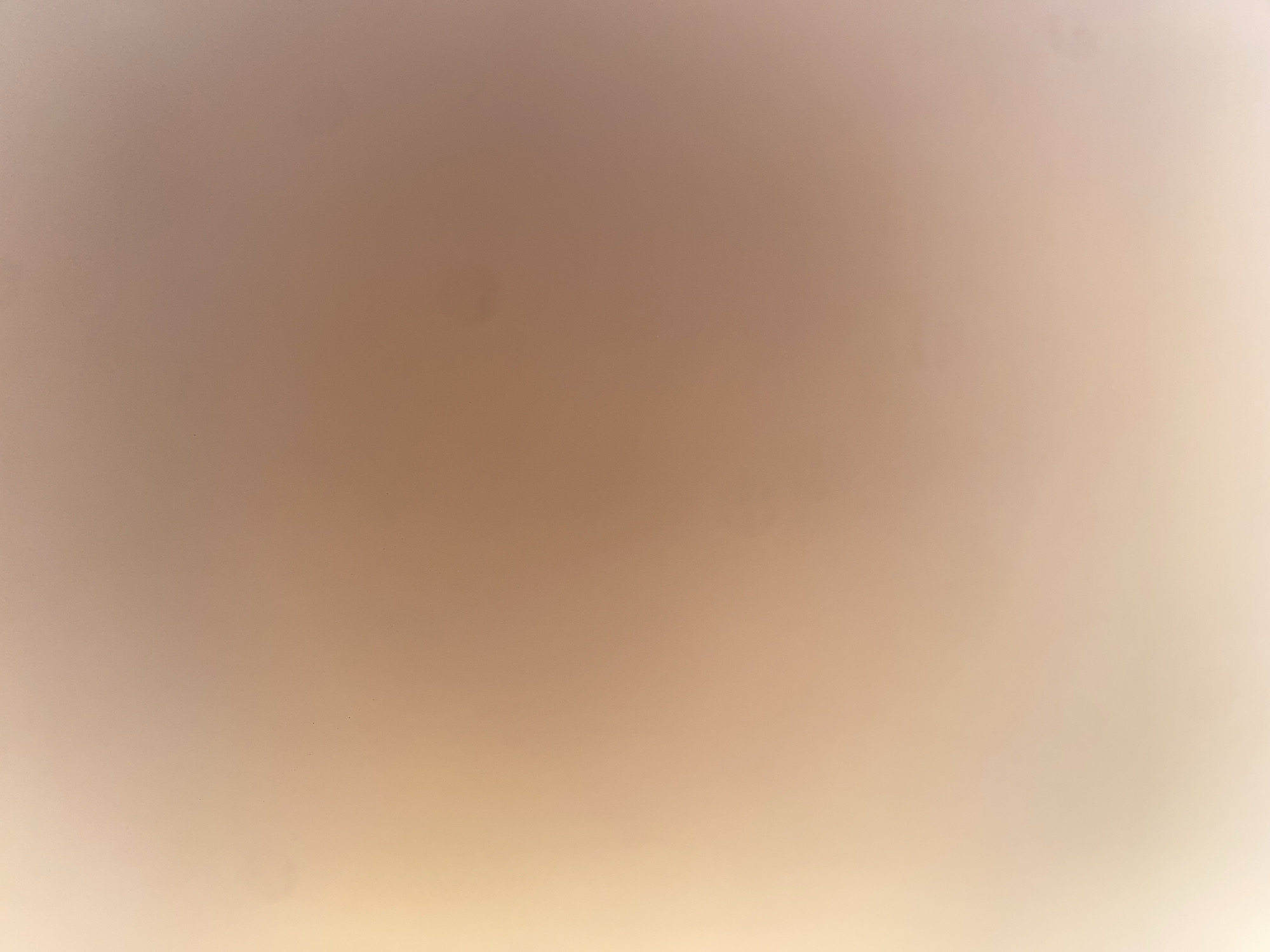 The image is a close-up, abstract shot of a textured surface, likely a wall or ceiling. It is uniformly lit with soft, warm light, creating a gradient effect from a bright, yellowish-white on the left side to a muted peachy-brown on the right. There are subtle variations in tone and some minor imperfections visible as small, dark specks scattered across the surface. No identifiable objects, people, or specific features of the environment are discernible. The scene is devoid of any context that would place it in Ablanitsa, Bulgaria, or any other specific location.