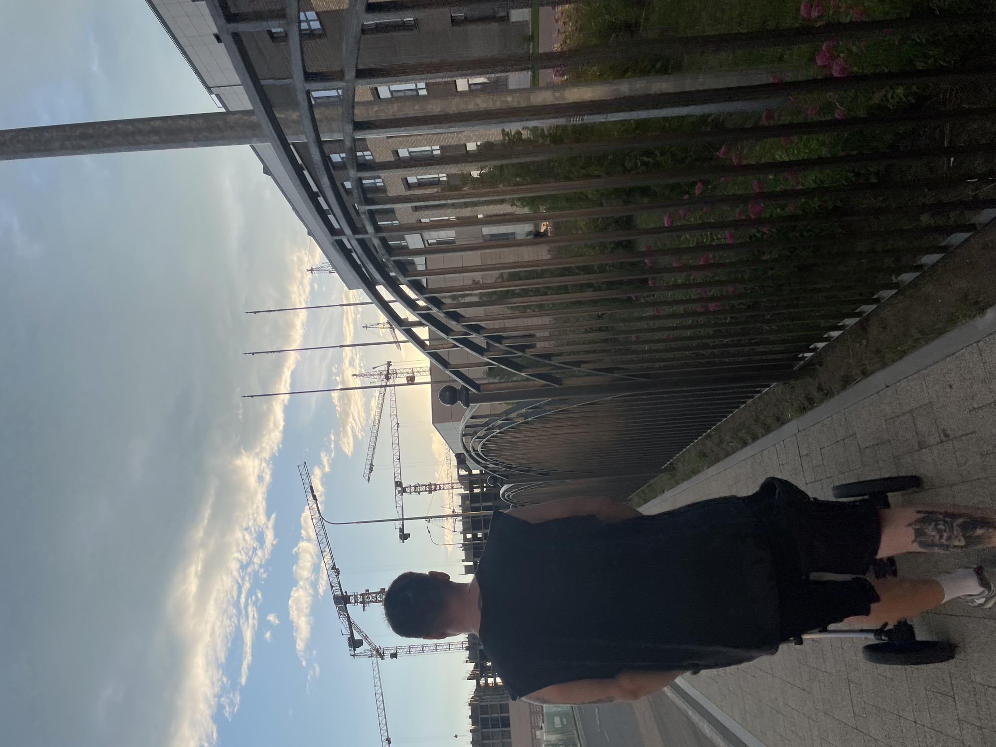 The image depicts a man from the back, pushing a stroller on a sidewalk. He is wearing a black sleeveless shirt and black shorts. Construction cranes are visible in the background, suggesting an ongoing construction site. A modern building with multiple windows is also present. The scene appears to be in an urban environment, possibly a residential area under development. The sky is partly cloudy, with hints of sunset colors. The location is identified as Stavropol, Russia, based on the prompt. The fence bordering the sidewalk has a decorative, curved design. There is greenery and small pink flowers visible behind the fence. The man's left leg has a tattoo.