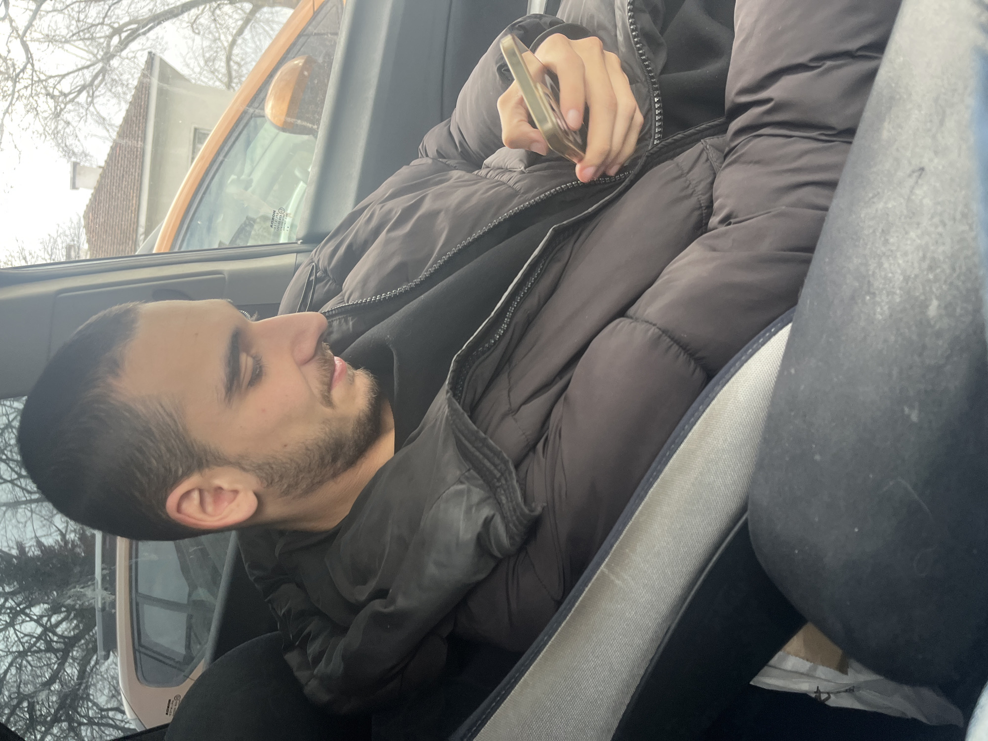 A young man is seated in the driver's seat of a vehicle, facing to the left and looking down at a cell phone held in his right hand. He has short dark hair, a beard, and is wearing a black puffer jacket. The vehicle appears to be parked or stationary, as an orange car and a building are visible through the driver's side window. Bare trees are visible in the background, suggesting a colder season, and based on visual clues the location is most likely in Varna, Bulgaria.