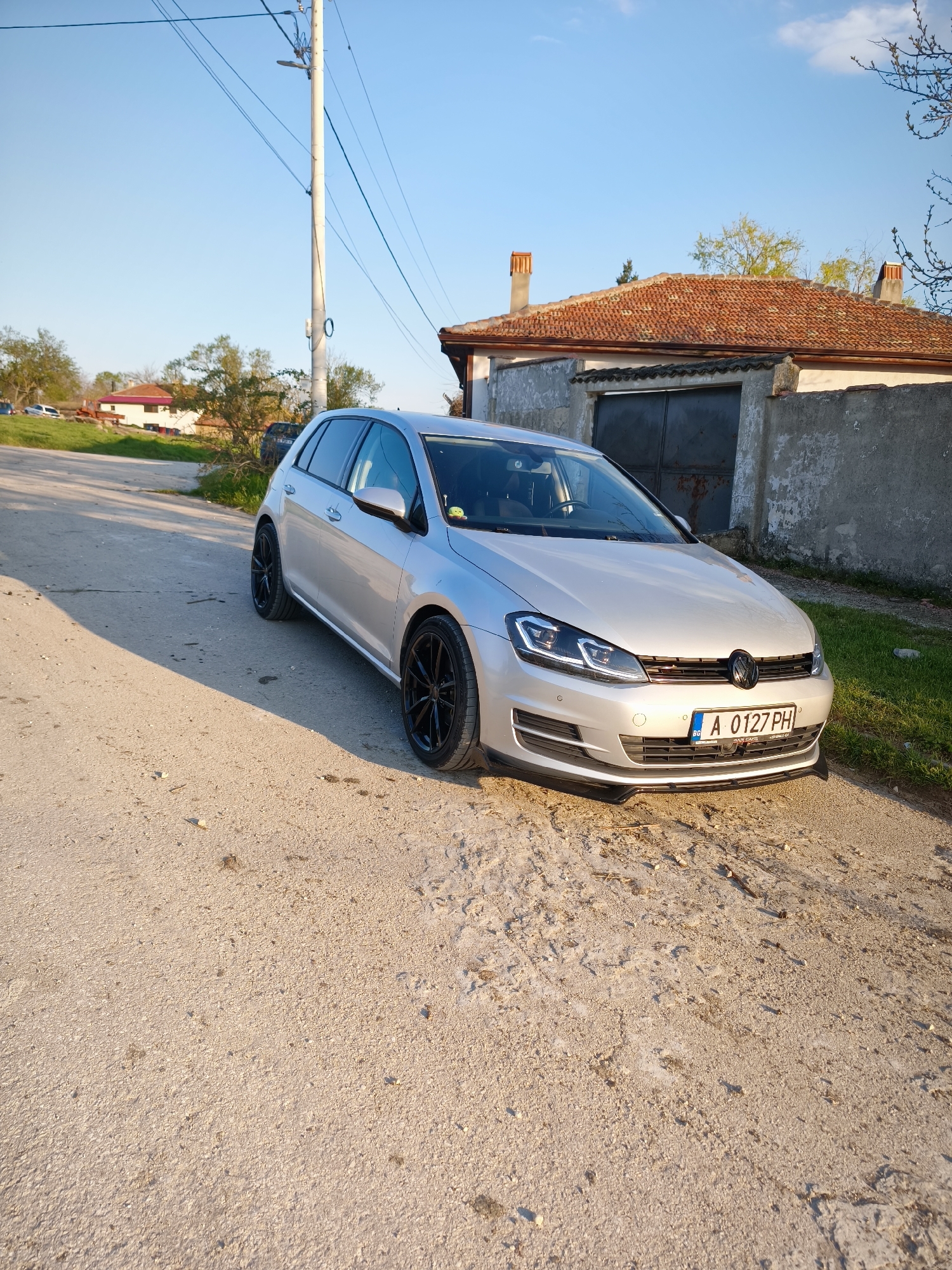 A silver Volkswagen Golf is parked on a cracked, light-colored road in front of a low, concrete building with a red tile roof. The building has a dark metal door. A utility pole stands to the left of the car. In the background, other houses are visible, and there are trees and a blue sky with some clouds. The car's license plate is visible and reads "A 0127 PH" with "BG" on the left side. The location is in Avren, Bulgaria. The scene is illuminated by bright daylight.