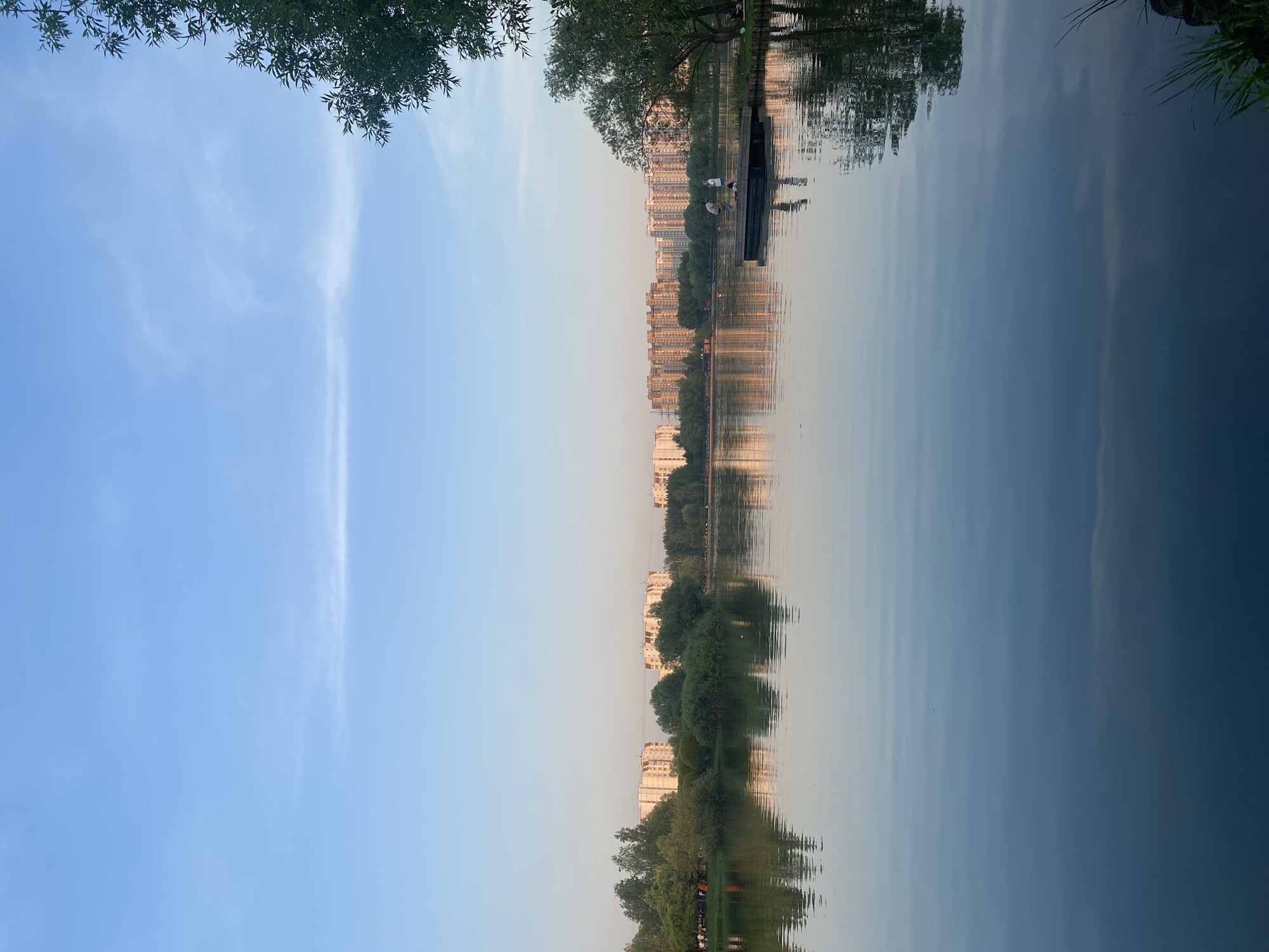 The image depicts a body of water, likely a lake or river, with reflections of buildings and trees creating a symmetrical composition. The sky is light blue with some cloud cover, and the water surface shows slight ripples. In the top right corner, a small wooden structure or pier extends into the water. Two individuals are visible on the structure. Given the architecture and environment, the location is possibly within the city of Moscow, Russia. The time of day appears to be dusk or dawn, suggested by the soft lighting.