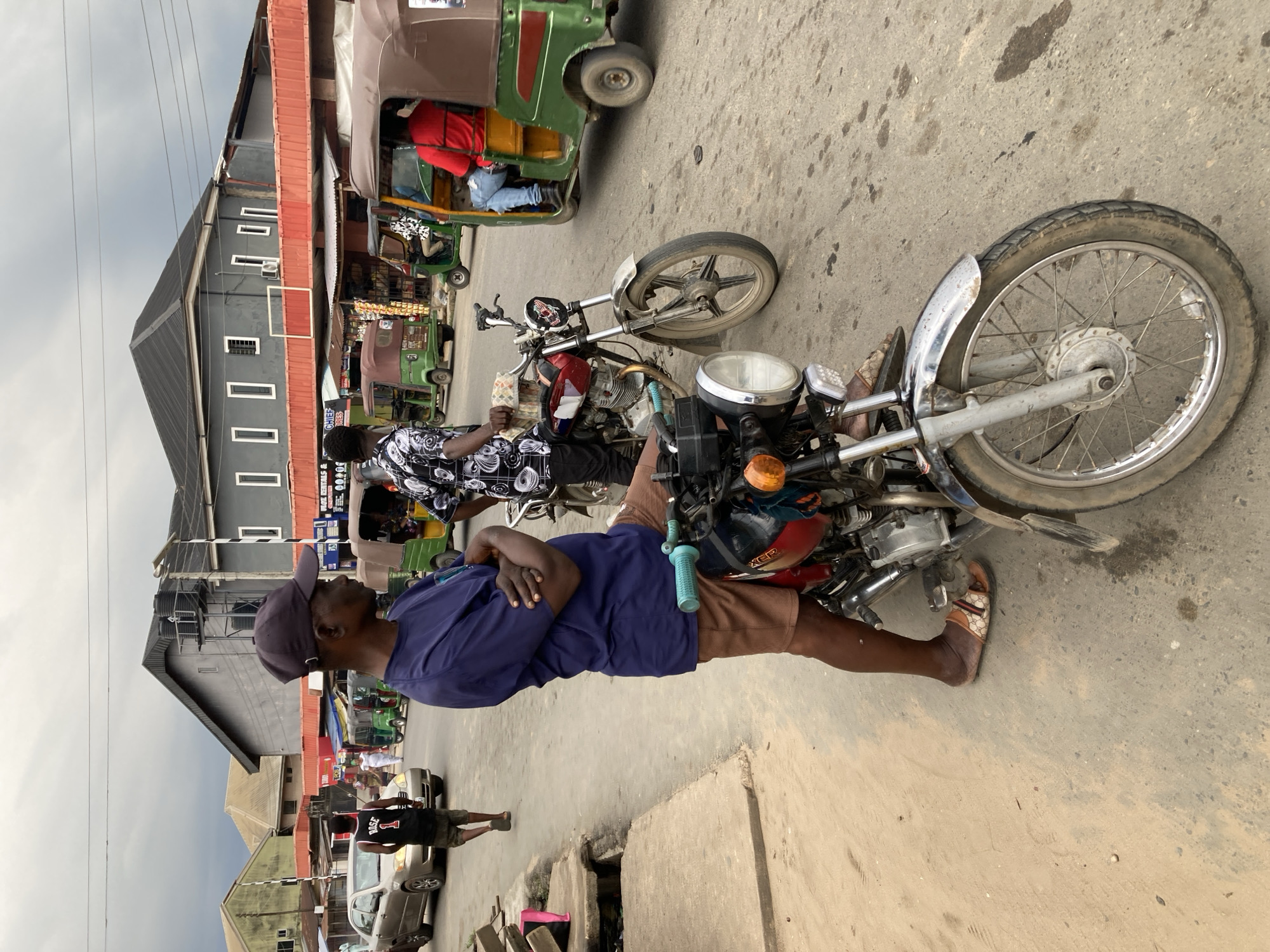 The image shows a street scene in Aba, Nigeria, with two motorcycles parked on the side of the road. A man in a blue shirt and brown shorts stands near one of the motorcycles with his arms crossed. Another man, wearing a black and white patterned shirt, stands near the other motorcycle. A tricycle is visible in the background. Other people and vehicles are present in the street scene. Buildings line the street, including one with a gray facade and a red awning. Power lines are strung across the sky. The street is paved and shows signs of wear and staining. A storm drain is located near the foreground.