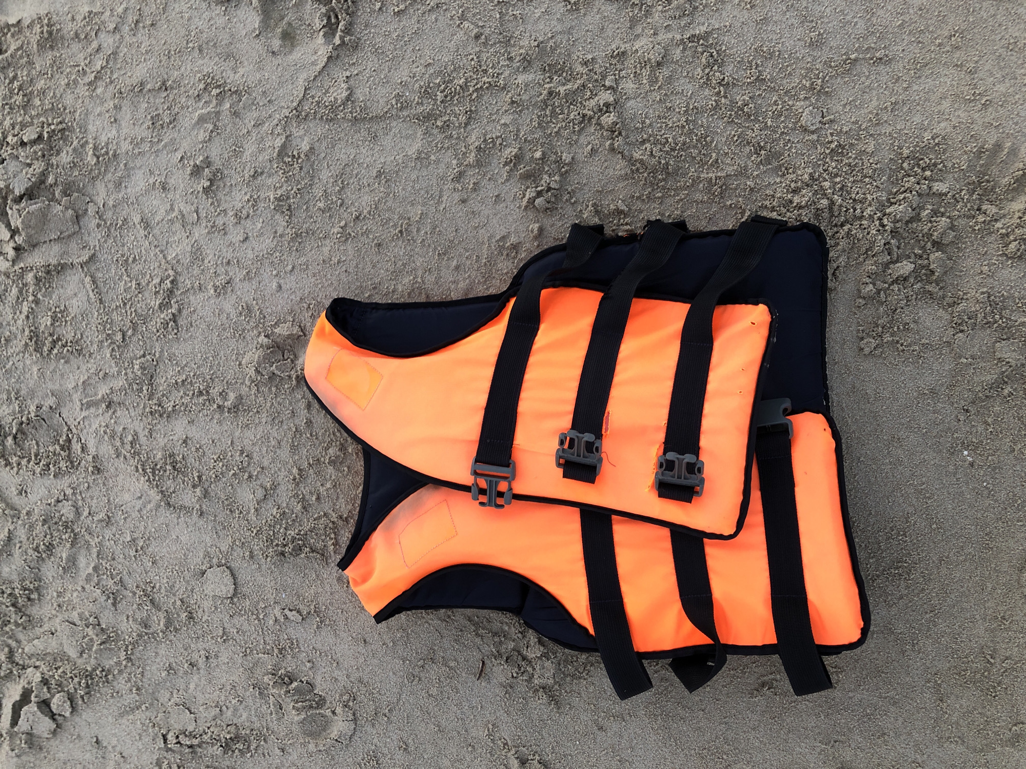 A bright orange life jacket with black straps and buckles lies on a sandy beach. The sand is a light gray color and appears slightly damp, with some textured patterns and indentations from footprints. The life jacket is folded slightly and positioned in the lower left quadrant of the image. The background is entirely sand, suggesting an outdoor environment, likely a beach in Đà Nẵng, Vietnam, given the typical context for such an item. No people are visible in the frame.