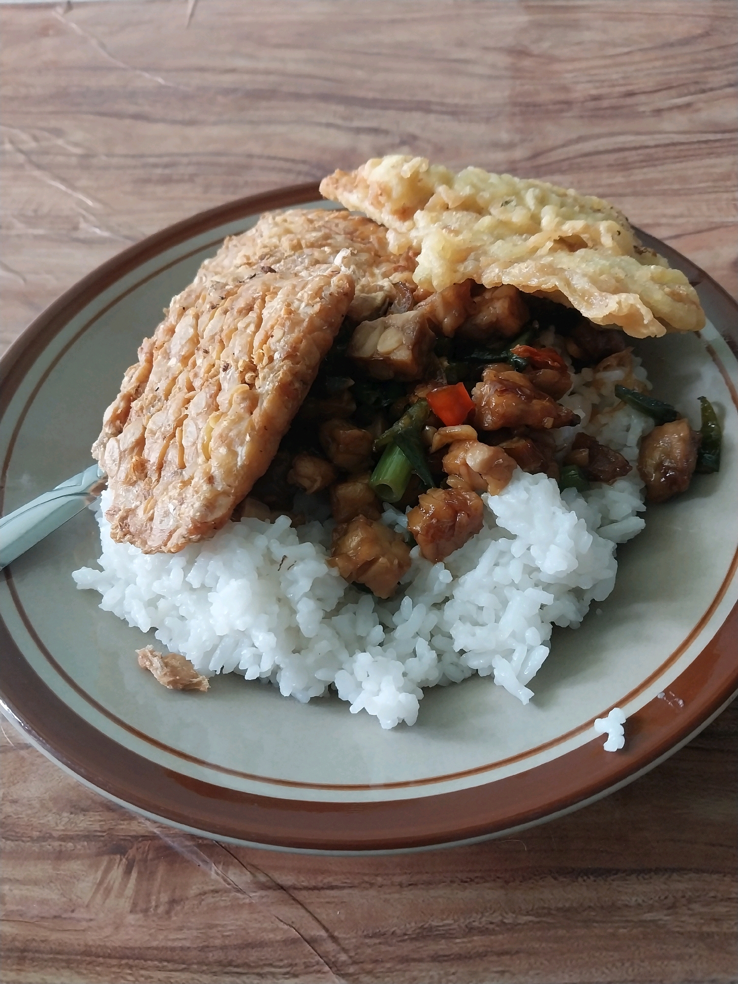 A plate of food is displayed on a wooden table. The plate contains white rice topped with a stir-fry of diced meat, green onions, and chili peppers, and is garnished with two pieces of fried tempeh. A fork rests on the left side of the plate. The setting appears to be indoors, likely in a restaurant or home in Wates, Indonesia.