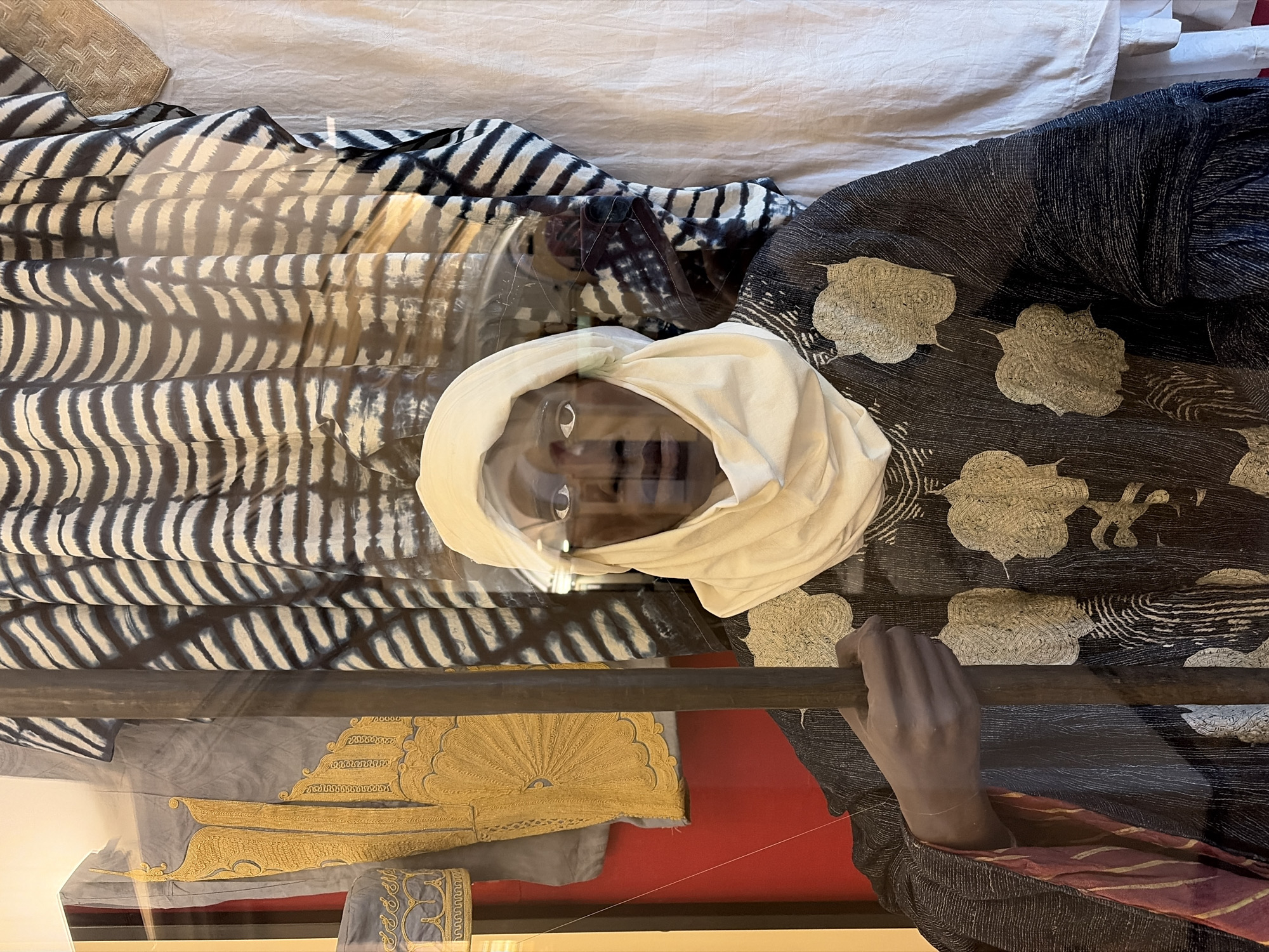 The image depicts a museum display, likely in Saint Petersburg, Russia, though this is not explicitly stated in the image. The display features a dark-skinned mannequin wearing a white turban-like head covering. The mannequin is partially visible behind what appears to be a glass case. The mannequin is dressed in a dark-colored robe with light-colored, stylized flower-like designs. One of its hands, also dark-skinned, grips a dark rod or staff. A portion of a blue garment with gold embroidery is also visible, as well as patterned fabrics and a white cloth background. The display appears to be illuminated by artificial lighting within the museum setting. The angle of the photograph is slightly overhead.