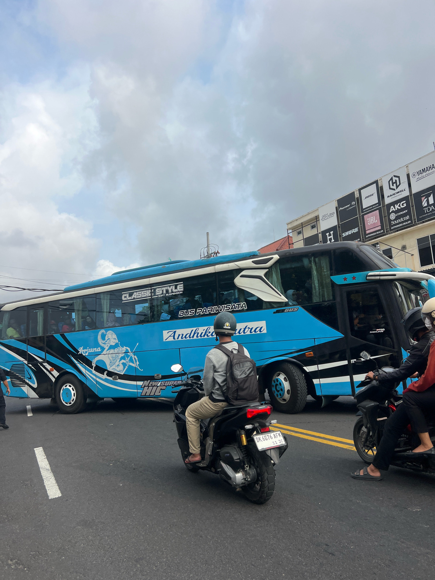 This is a medium shot, taken from a slightly elevated angle, of a busy street scene in Denpasar, Indonesia, with a large blue and black tour bus dominating the frame. The bus is adorned with a white graphic of a mythological figure on its side, and text that reads "CLASSIC STYLE" and "BUS PARIWISATA". Several people are visible inside the bus through the tinted windows.
In the foreground, two motorcycles are stopped at what appears to be a traffic light. The rider of the front motorcycle, a male wearing a backpack and a helmet, is seen from behind. The motorcycle behind him has two occupants, a male driver and a female passenger in the back. The female passenger's legs and sandals are visible. The motorcycles are stopped on an asphalt road with visible white and yellow lane markings.
In the background, a building with a facade displaying various brand logos such as "YAMAHA", "BOSE", "AKG", "JBL", and "SHURE" is visible. The sky is overcast with a mix of grey and white clouds. The overall impression is a typical urban traffic scene in Denpasar during daylight hours.