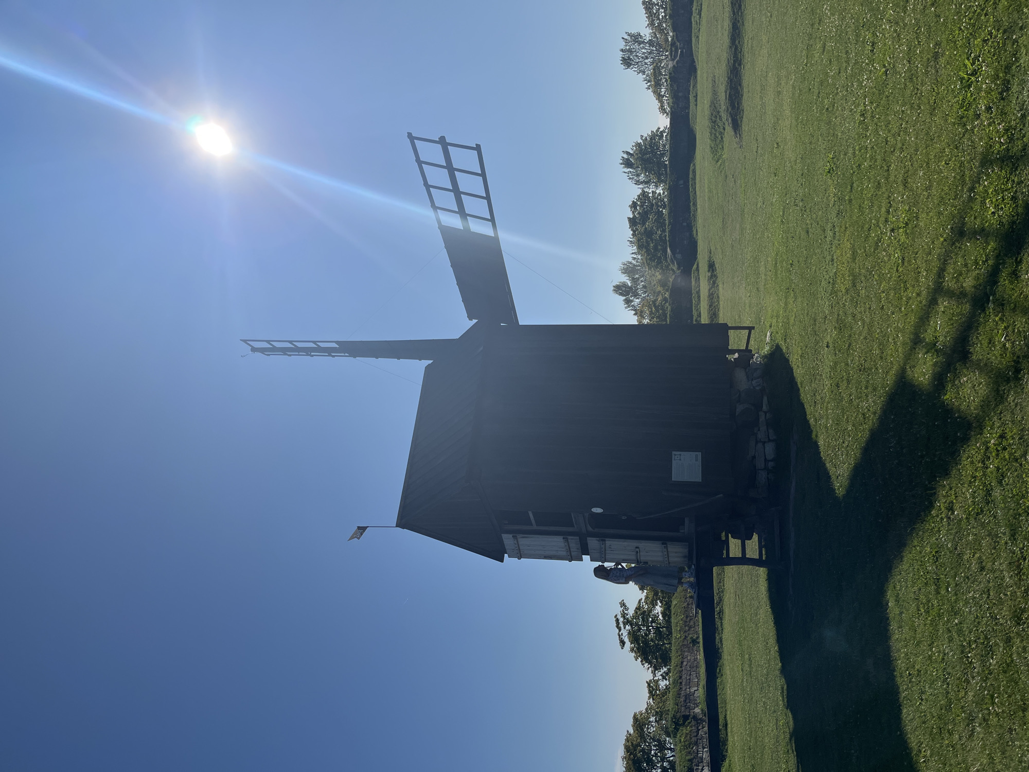 An outdoor, full-shot image depicts a historic, dark wooden windmill under a clear, bright blue sky with the sun prominently featured in the upper right quadrant, casting a strong lens flare. The windmill is positioned slightly left of center, with its large, weathered blades visible against the sky. A person, appearing as a silhouette, stands on a wooden platform attached to the side of the windmill, next to an open doorway. The windmill is situated in a grassy field, with a low, stone wall and trees visible in the background, suggesting a historical or park-like setting. The ground is covered in green grass with some undulations. The overall scene is bathed in bright sunlight, creating sharp shadows. This windmill is located at the Kuressaare Episcopal Castle in Kuressaare, Estonia, a preserved medieval fortress.