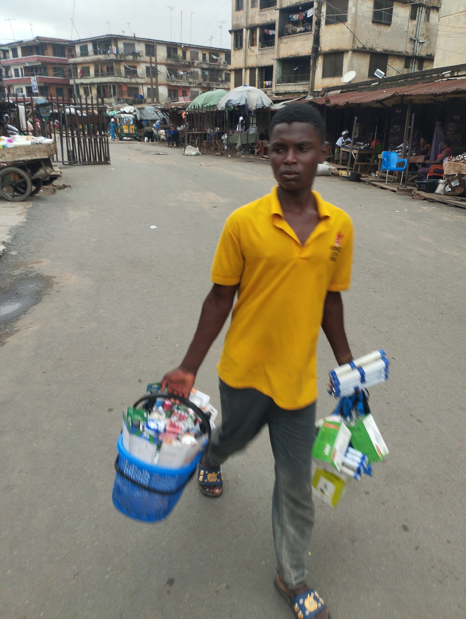 A young man in a yellow polo shirt and gray pants walks down a gray asphalt road in Fegge, Nigeria. He carries a blue plastic basket overflowing with miscellaneous items in his left hand and a bundle of packaged goods in his right. The background shows a bustling street market scene with buildings, stalls, and people. The sky is overcast.