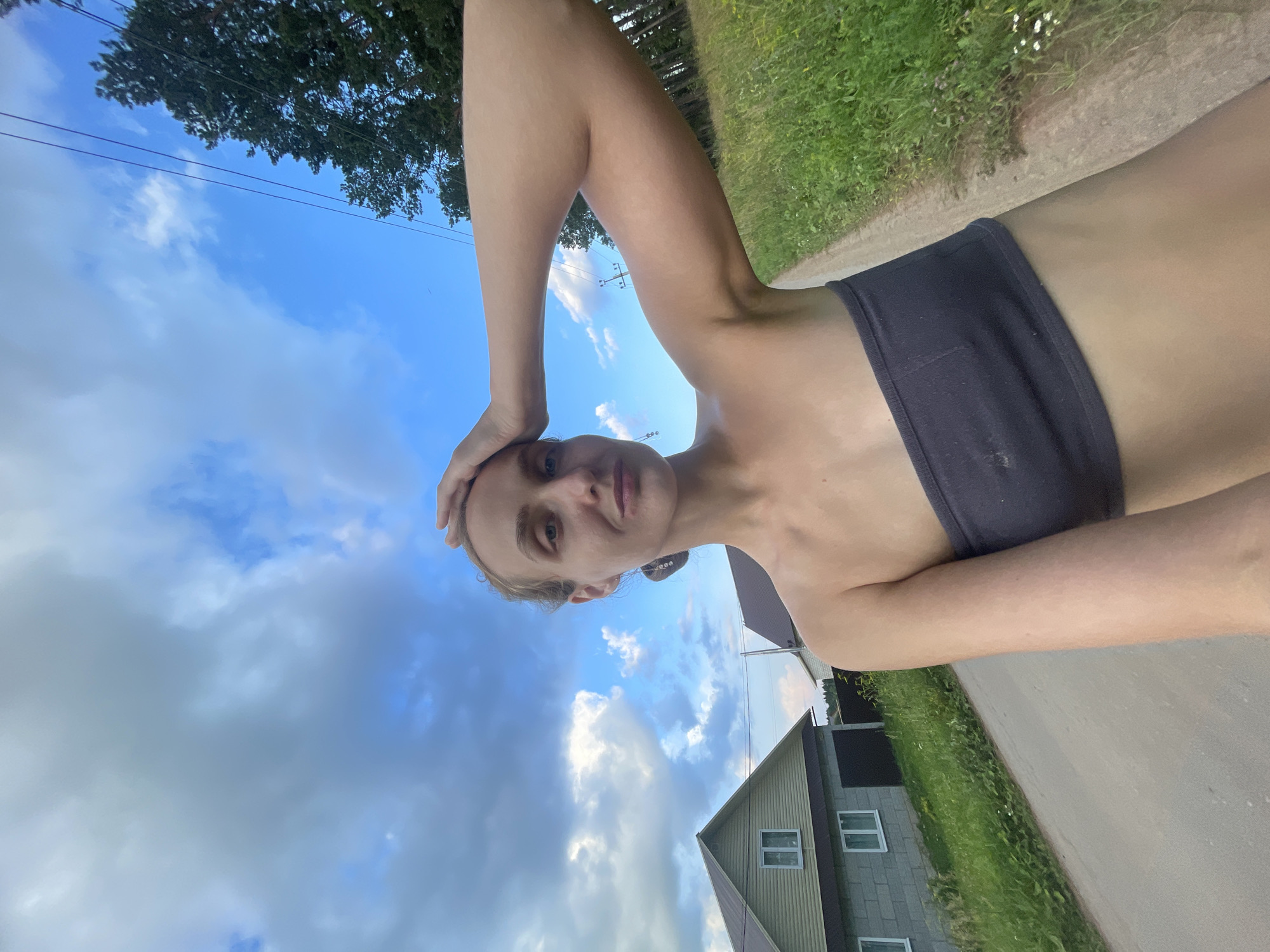 An outdoor, waist-up, selfie-style photograph captures a woman with blonde hair pulled back, wearing a dark grey tube top. She is standing on an asphalt road, with her right arm raised and her hand resting on her forehead. To her left, a light green house with a grey foundation and white-trimmed windows is visible, alongside a smaller, darker-roofed building. Lush green grass and wildflowers border the road. In the background, a line of trees stands against a bright blue sky dotted with fluffy white clouds. Electrical wires run diagonally across the upper right portion of the frame. The scene appears to be set in a rural or suburban area, potentially in the village of Podshivalovo, Russia.