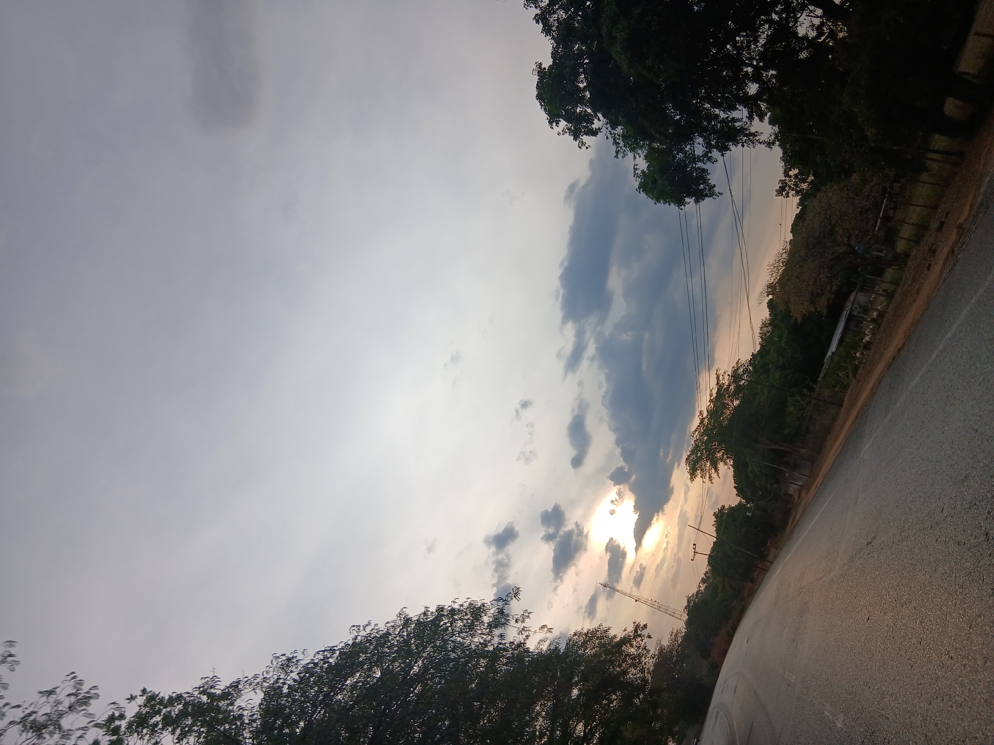 The image shows a view of Lomas del Viento, Venezuela, facing an asphalt road with trees lining the side and the sky above. The sky is overcast with a mix of clouds and visible sunlight. Electrical wires and poles run along the tree line. The photograph is taken at an angle, causing the horizon to be tilted.