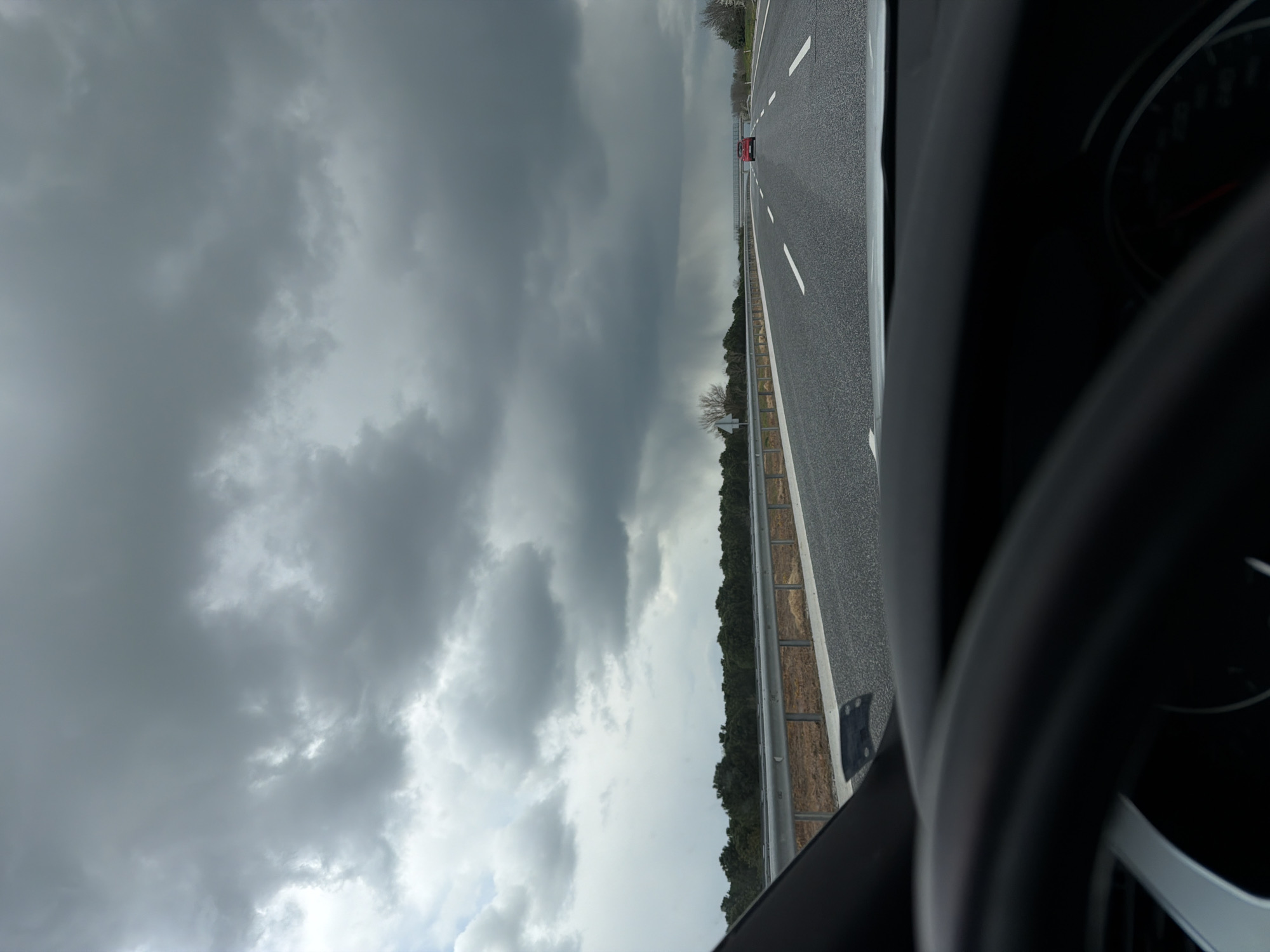 The image shows a highway scene, taken from the interior of a moving car. A red car is seen further ahead on the road. The sky is overcast with heavy, gray clouds. The road has white lane markings and a guardrail runs along the side, separating the highway from a wooded area. The location is likely near Havsa, Turkey, given the provided context. The steering wheel and dashboard of the car are partially visible in the foreground.