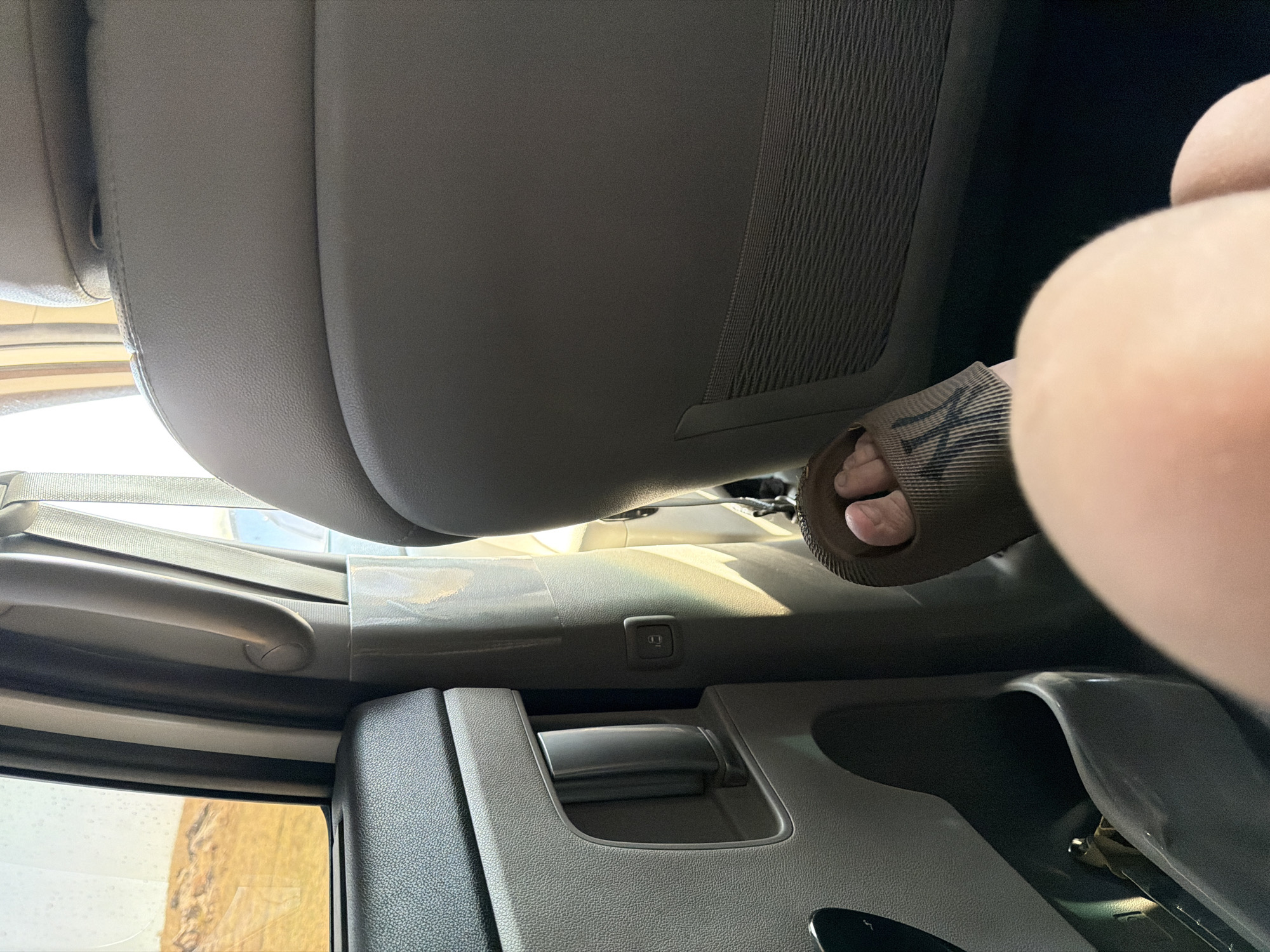 A person's foot wearing a brown slide sandal with a tattoo on the instep is visible in the foreground, resting on the floor of a vehicle's interior. The sandal appears to have a ribbed texture. The person's leg and knee are also in view, with smooth, light-toned skin. The background shows the interior of a car, including a grey plastic door panel with a storage compartment and a silver handle, a beige seat, and a window offering a view of a dry, arid landscape with hills. Sunlight streams through the window, casting shadows and highlighting the textures of the car's interior. A small, dark-colored object, possibly a pen or stylus, is visible inside the door's storage compartment. The overall scene suggests travel. The image does not provide specific information about the location within the city of Теректі, Kazakhstan, beyond it being a vehicle interior.