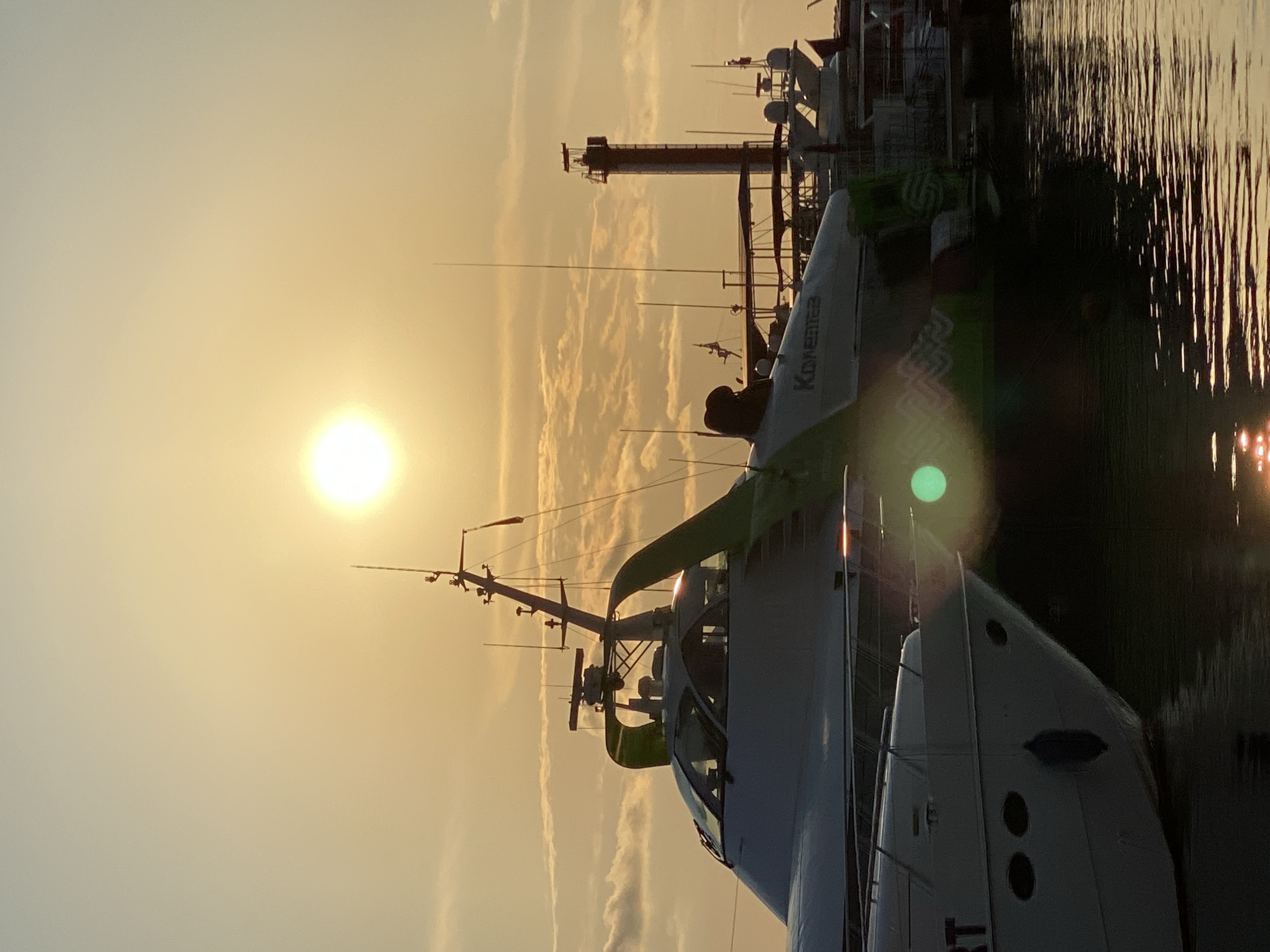 The image, oriented vertically, depicts a harbor scene at sunset in Sochi, Russia. The sun, a bright orb, is low in the sky, casting a warm, golden glow across the clouds and the water. The sky is filled with wispy, elongated clouds that are illuminated by the setting sun, creating a dramatic and serene atmosphere.
In the foreground, the stern of a white boat is visible, with several dark, oval portholes. Part of a green hull is also visible on the left side of the boat. The boat appears to be docked alongside other vessels and structures in the harbor.
The background features a collection of masts, antennas, and the silhouettes of other boats and port infrastructure, including what looks like a tall, red and white tower with a cylindrical structure at the top, possibly a lighthouse or a radio tower. The water reflects the colors of the sunset and the surrounding structures, creating shimmering patterns.
The overall impression is one of a peaceful evening in a coastal city. The time of day is clearly sunset, with the warm light and long shadows. The weather appears clear with scattered clouds. The image does not show any people. Visible text on the white boat reads "KONISHTER".