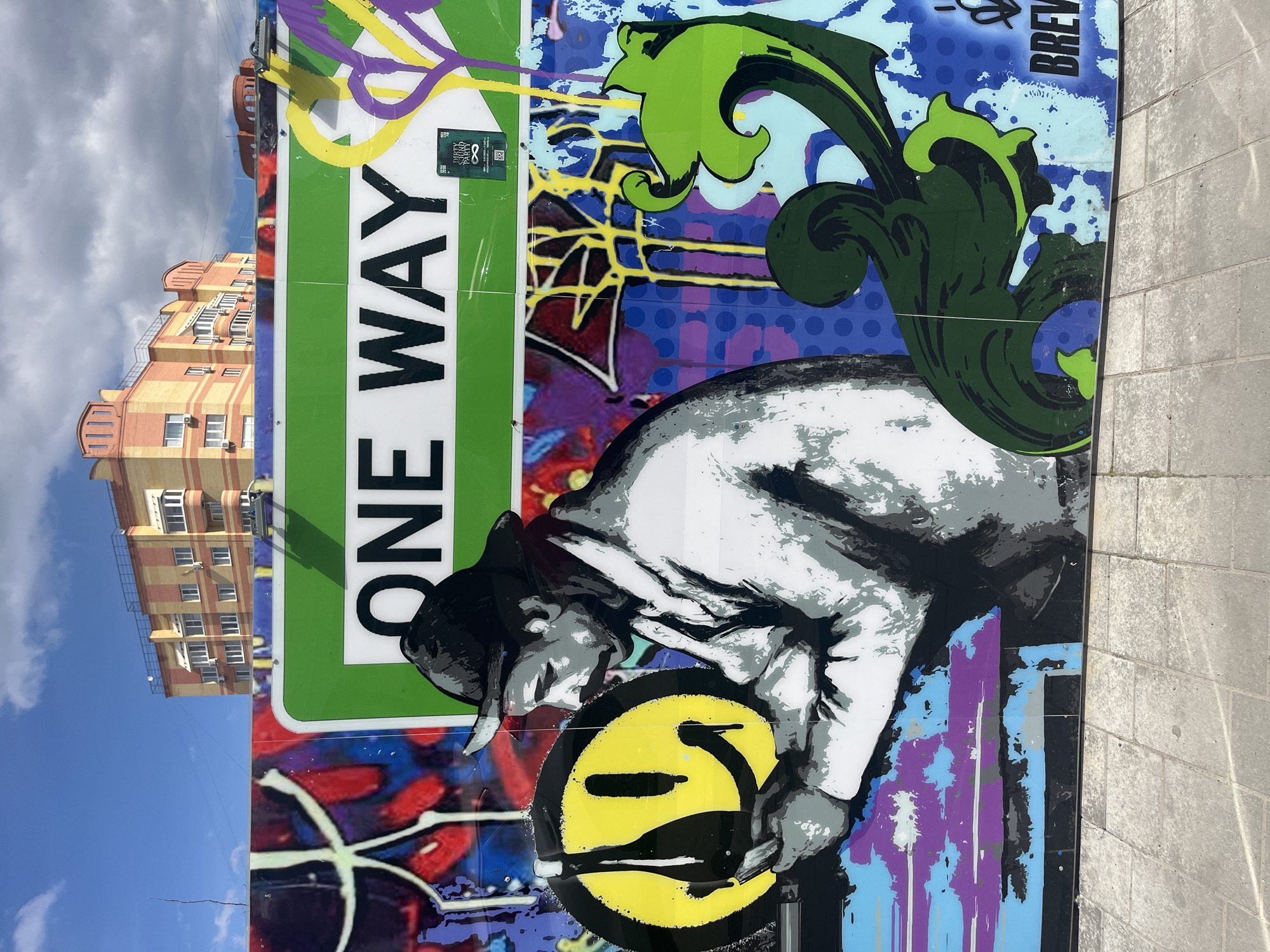The image shows a vibrant mural on a wall in Ulyanovsk, Russia. The mural features a large, vertical "ONE WAY" street sign with green background and white lettering. Adjacent to the sign is a monochrome depiction of a person wearing a hat and coat, bent over and seemingly working on a yellow smiley face. The smiley face is partially obscured by a black object that appears to be a bottle or a tool.
The background of the mural includes various graffiti-style elements in different colors, such as purple, blue, yellow, and red. A building with yellow and orange brick walls and multiple windows is visible in the background, along with a blue sky with scattered clouds. The mural is placed on a gray brick or concrete wall.