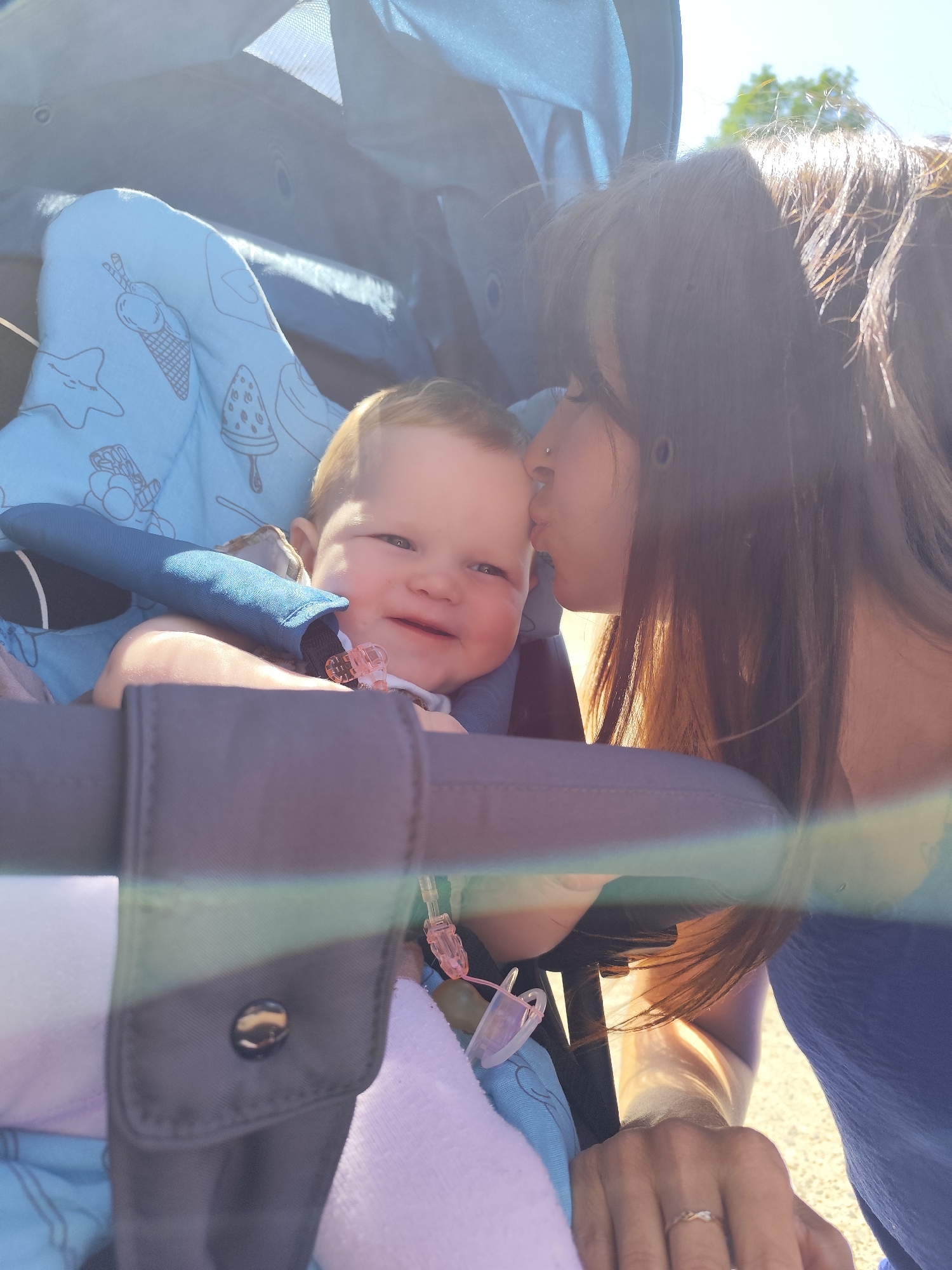 A woman is kissing a baby on the forehead. The baby is in a stroller with a blue patterned liner. The woman has long dark hair and is wearing a blue top. She is holding onto the stroller. The photo appears to be taken outdoors in bright sunlight. The exact location in the city of Фоки, Russia cannot be determined from the image.
