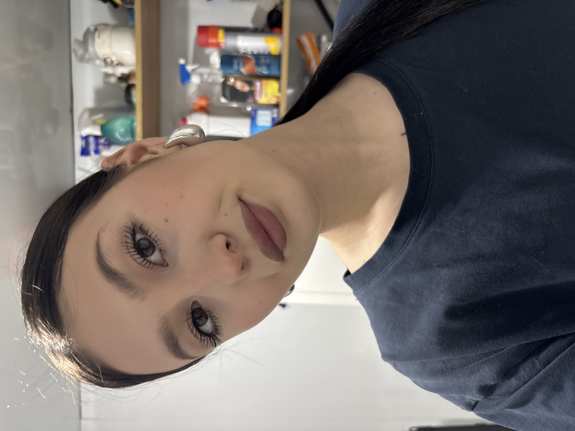 A woman with brown hair and silver earrings is depicted indoors. She is wearing a dark-colored shirt. The background includes shelves with various household items. It is not possible to determine the exact location in the city of Фоки, Russia, based on the given image.
