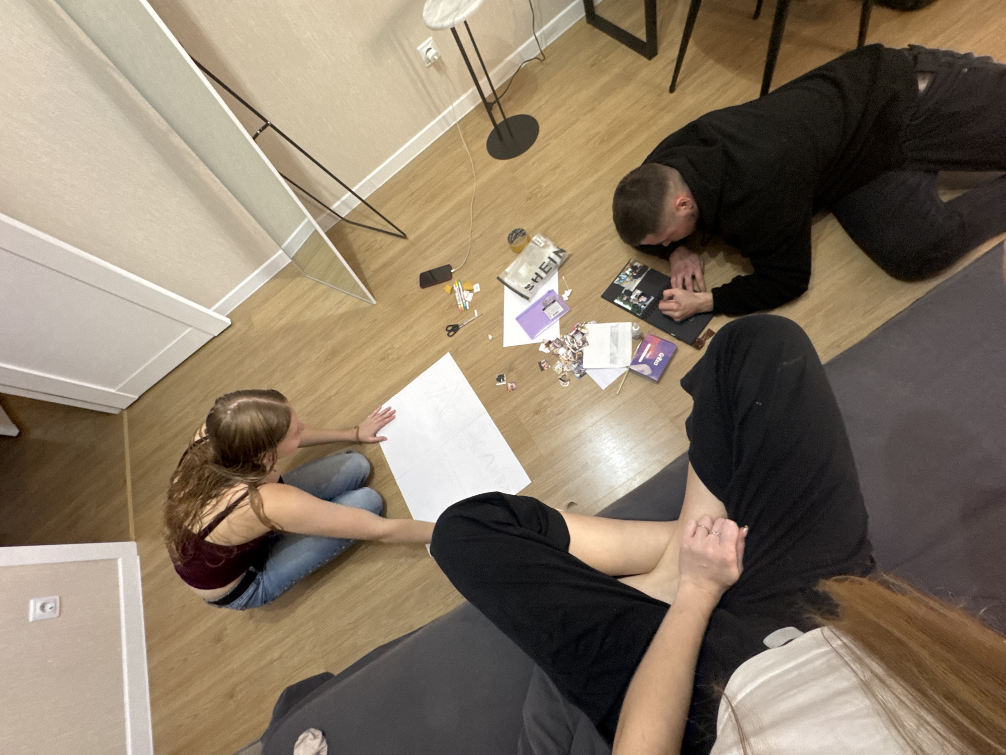 The image depicts an indoor scene, likely an apartment living room with light-colored walls and a wooden floor. The room contains a large mirror, a floor lamp, and a sofa. Three individuals are present: one person is kneeling on the floor, seemingly engaged with a large sheet of white paper. Another person is lying on their stomach, interacting with a laptop, while a third person's legs are visible on the sofa, suggesting they are also in the room. Various items are scattered on the floor, including a mobile phone, scissors, pens, a pouch with the "SHEIN" logo, and small printed images or stickers. The overall impression is one of casual activity, possibly creative or collaborative, taking place in a domestic setting. There is no indication of the location being in the city of Perm, Russia, or any other specific city.