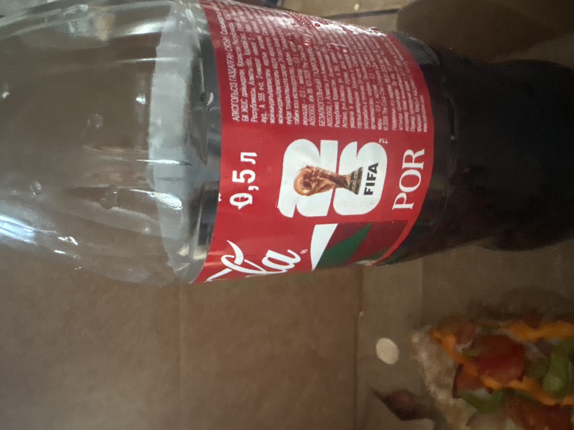 The image shows a close-up of a Coca-Cola bottle with a red label indicating it is a 0.5L size. The label features the FIFA World Cup trophy and the word "FIFA" in white text. Part of a pizza slice is visible in the lower left corner. The text on the bottle appears to be in Russian or a similar Cyrillic-based language, with some of it relating to production and ingredients. While the image itself doesn't directly confirm the location, if this bottle was purchased in Aktobe, Kazakhstan, it would be a common consumer product found in households or retail establishments within the city. The overall environment appears to be indoors, with a neutral brown background.