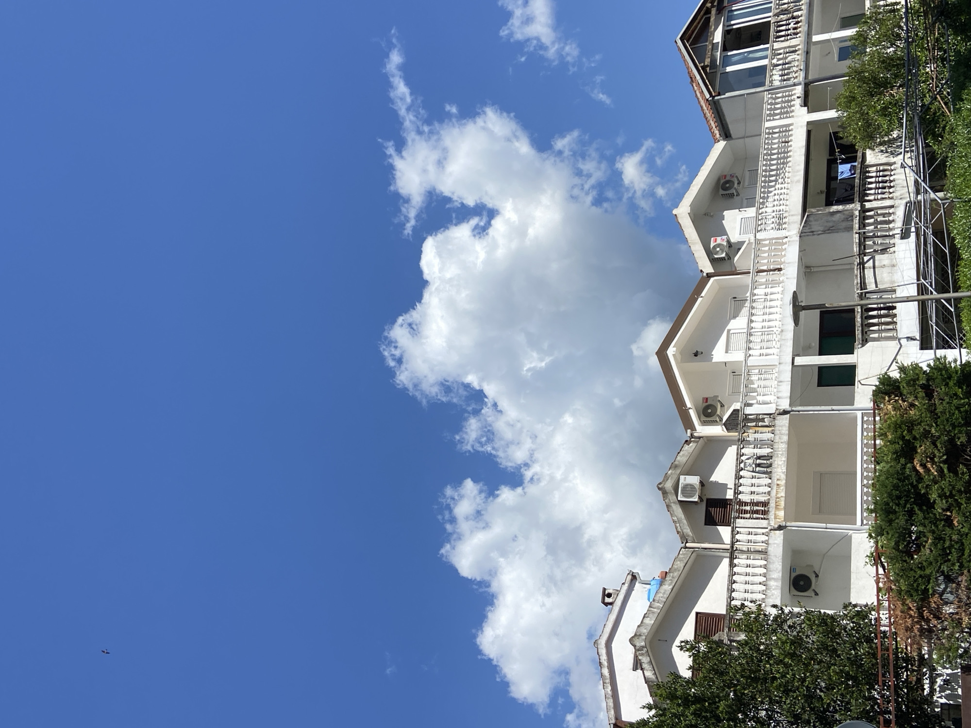 The image shows a multi-story white building against a bright blue sky with cumulus clouds. The building has a series of balconies with white balustrades, and air conditioning units are mounted on the exterior walls of each level. Trees are visible to the right of the building, partially obscuring the lower floors. A small object, possibly an aircraft, is visible in the lower left portion of the sky. This image appears to be taken in Budva, Montenegro.