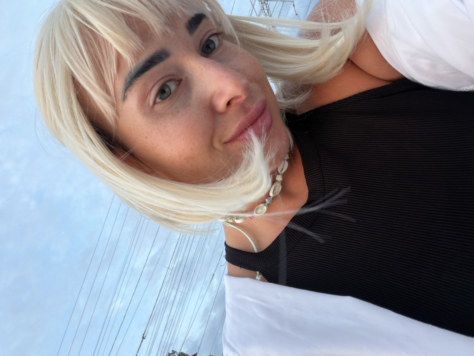 A medium shot captures a woman with blonde hair and a black top, posing for a selfie. The background reveals a cloudy sky with numerous power lines. The woman's head is tilted, and she is smiling at the camera. She is wearing a necklace made of seashells and beads. A white garment, possibly a jacket or shirt, is draped over her left shoulder and arm. The overall setting appears to be outdoors, possibly on a street or walkway in Sochi, Russia, given the visible electrical infrastructure commonly seen in urban environments.