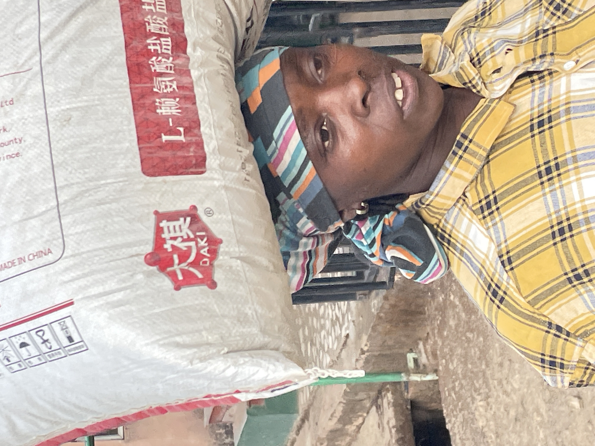 The image shows a person in Jos, Nigeria lying on their back next to a large white bag with red lettering in Chinese. The person is wearing a yellow plaid shirt and a striped headscarf. The background is a concrete surface with a green pipe.