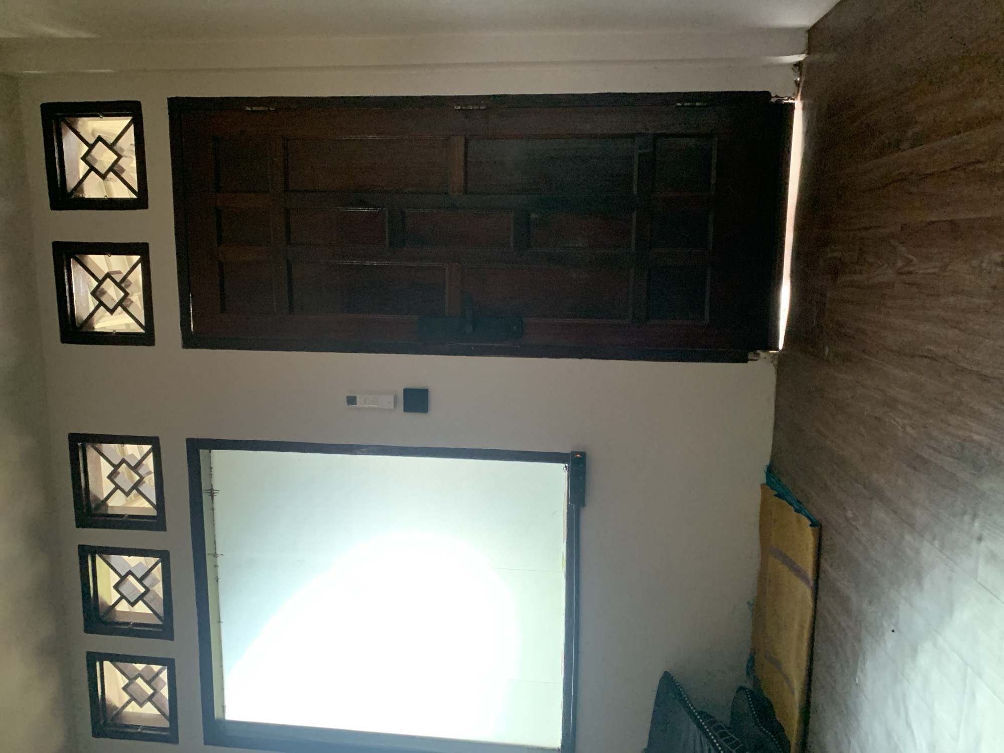 A low-angle interior shot captures a dark wooden door and a large frosted window, both set into a white wall. Above the window, three decorative square vents are visible, each with a diamond-shaped grill pattern. To the left of the door, a small, rectangular white device is mounted on the wall, possibly a light switch or remote control. Below the window, a yellow mat lies on the wooden floor, next to a dark object, perhaps a cushion or bag. The bottom of the door is slightly ajar, allowing a bright light to spill onto the floor. The scene appears to be located in Kualadua, Indonesia, based on the architectural style and the lighting conditions, which suggest a tropical climate.