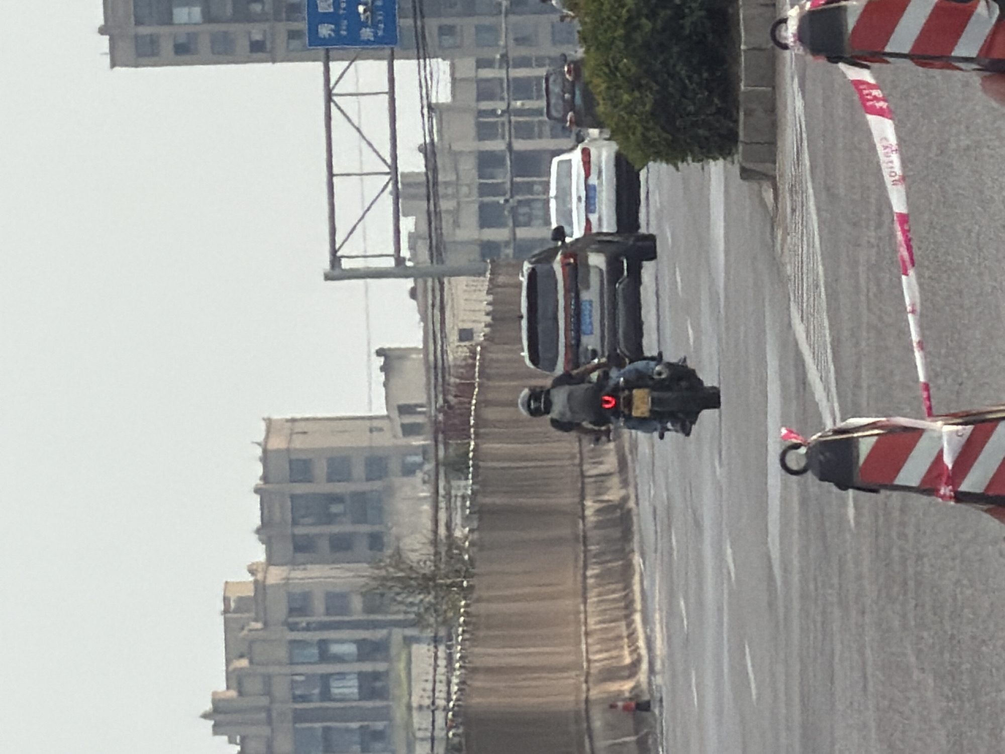 The image depicts a street scene in Jiangjin, China, with a focus on traffic. In the foreground, a motorcycle rider wearing a black helmet and backpack is moving away from the camera on the right lane of a multi-lane road. The motorcycle has a yellow license plate with visible characters and a red taillight. To the left of the motorcycle, a portion of a traffic cone with red and white diagonal stripes is visible. Further down the road, a silver SUV is in the left lane, followed by a white sedan, and then a dark colored SUV. In the background, several multi-story buildings line the street, with some appearing to be under construction or of older architecture. A brown barrier runs along the side of the road. A blue traffic sign with Chinese characters is mounted on an overhead gantry. The sky is overcast.