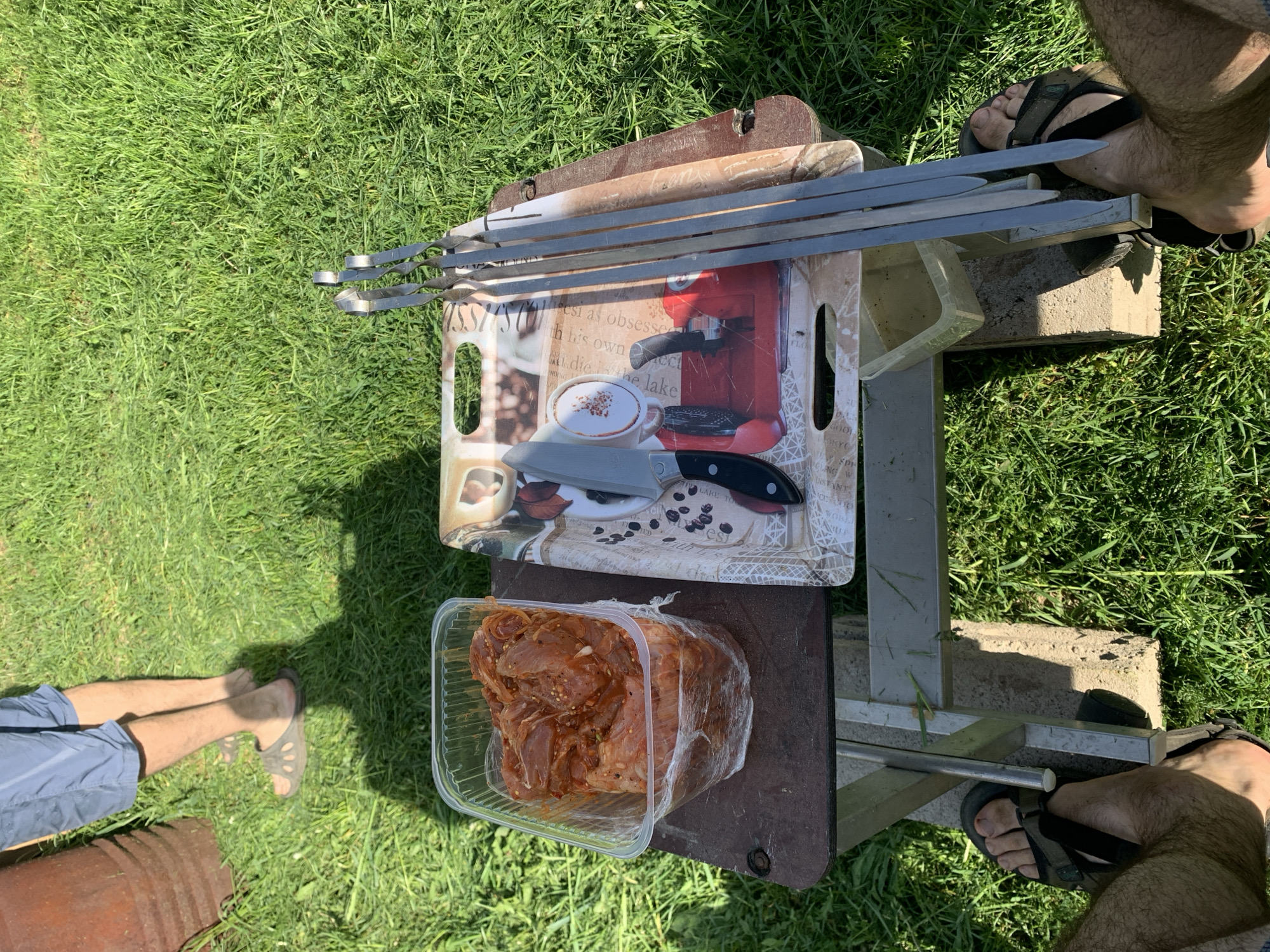 The image depicts an outdoor preparation scene for a barbecue, likely on a grassy area. Visible are skewers, a plastic container of marinated meat, a tray with a coffee-themed design holding a knife and another tool, and a makeshift table structure built from metal pieces and supported by concrete blocks. Parts of two individuals are visible in the foreground: one with legs in shorts and sandals to the left, and another with feet in sandals positioned to the right. The photo appears to be taken in daylight, casting shadows on the grass. Without specific information about the locale's attributes, it's difficult to pinpoint the precise location within Chaikovsky, Russia; however, given the scene's nature, it would likely be a backyard, park, or recreational area.