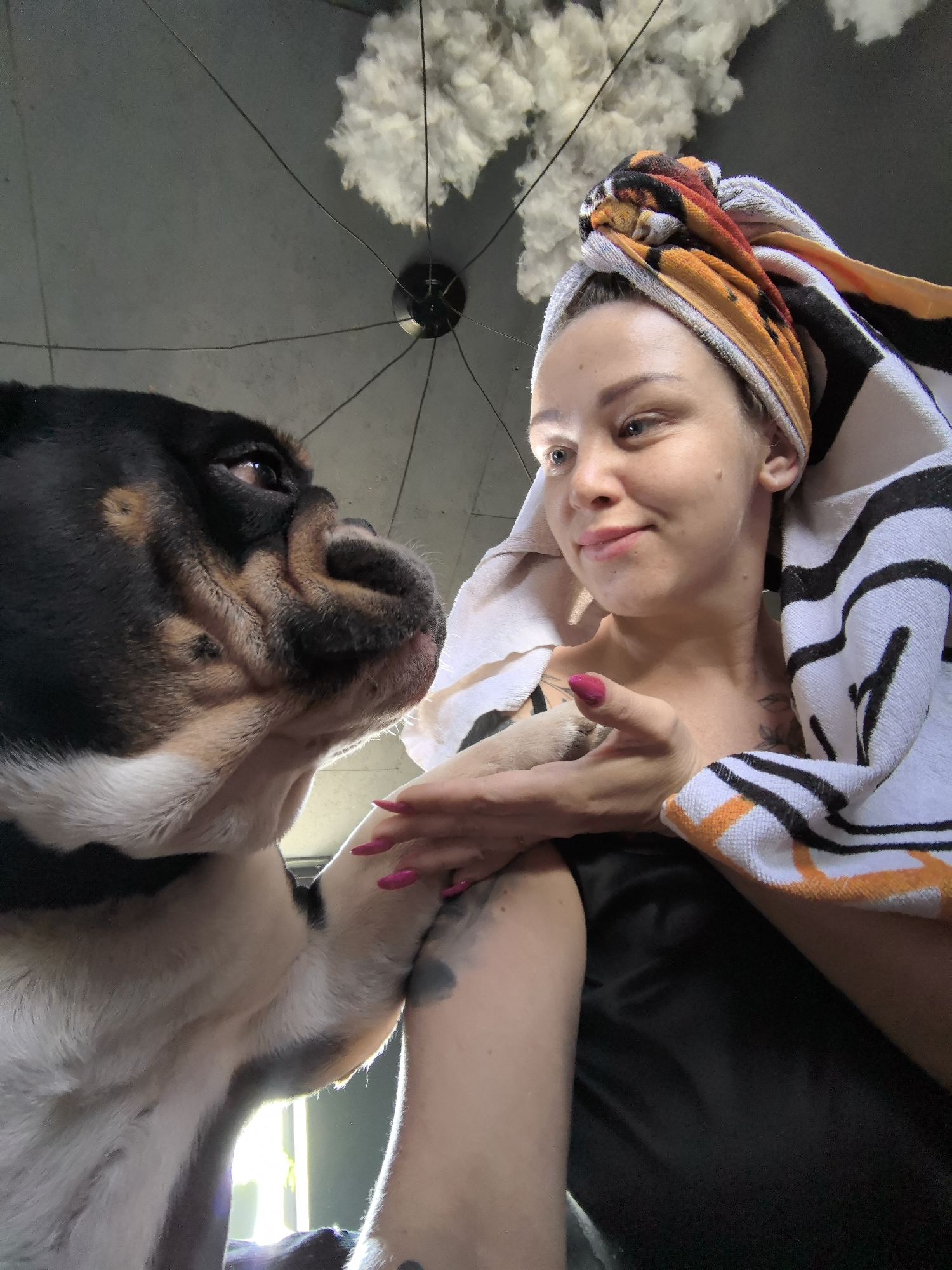 This close-up indoor photograph from Noviy, Russia, captures an intimate moment between a woman and a bulldog. The woman, shown from the chest up, has her hair wrapped in a colorful, striped towel (featuring orange, black, and white patterns), suggesting she may have recently showered. She smiles warmly, looking down towards the dog, her light blue eyes and pink-painted fingernails prominent. A dark tattoo is visible on her left arm, and she wears what appears to be a black top.
The bulldog, with its distinctive black, tan, and white fur and wrinkled face, is positioned on the left side of the frame, looking up intently at the woman. Its right paw rests gently on her arm, indicating a bond of affection.
In the background, a unique ceiling fixture is visible, featuring a fluffy, white, cloud-like element from which thin dark wires extend to a central point. The lighting is soft, suggesting a relaxed daytime atmosphere indoors. The overall activity depicts a heartwarming interaction and mutual affection between the woman and her dog. There is no visible text in the image.