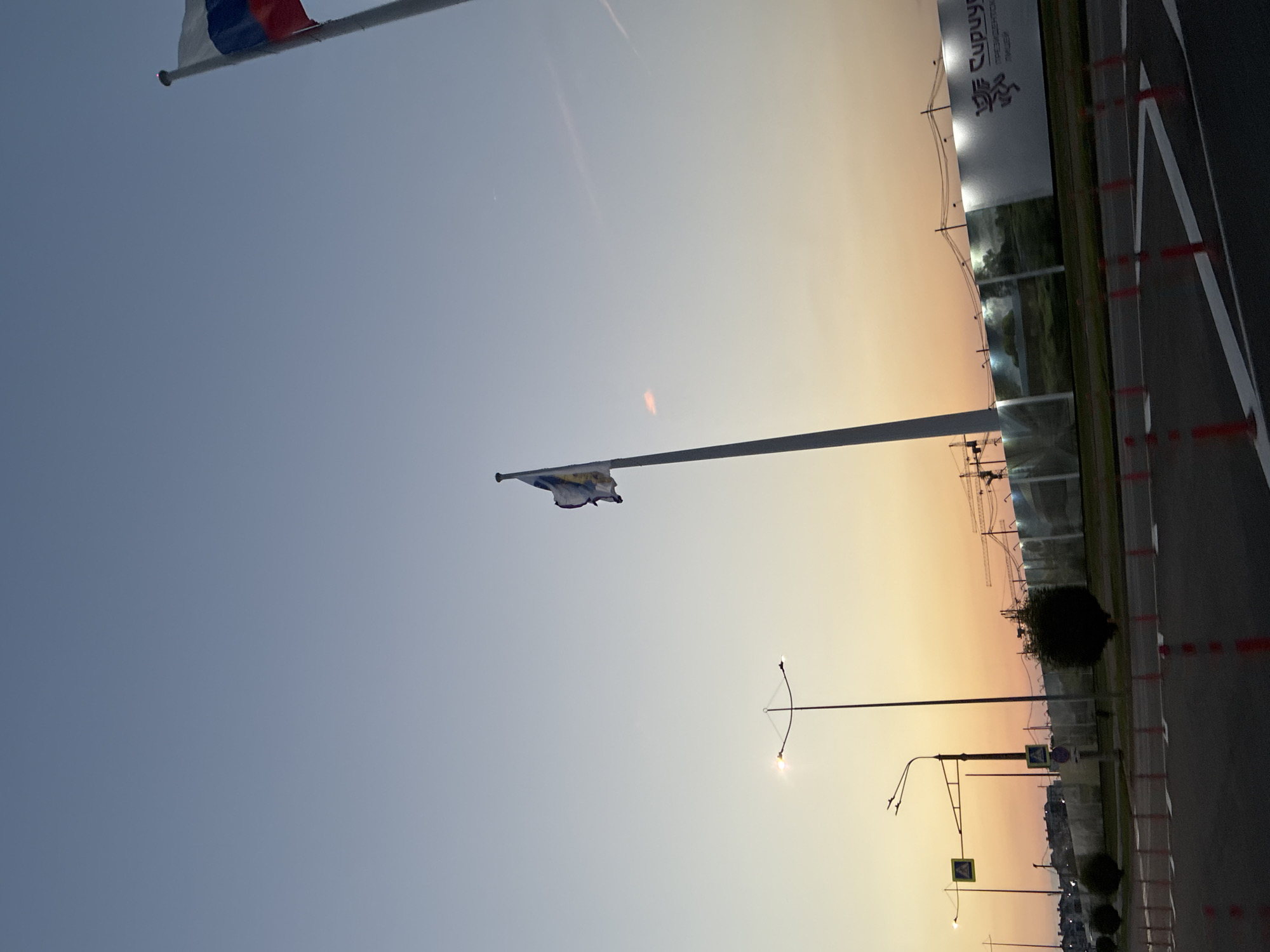 The image captures a dusky sky over a city street in Sirius, Russia. In the foreground, a road with white markings and red traffic cones is visible. A pedestrian crossing sign with a yellow diamond shape is mounted on a pole to the left, accompanied by streetlights. Further back, a construction site is partially obscured by a fence adorned with large images, one of which displays a stylized emblem and text in Russian, possibly indicating "Cupuyc" and "PRESIDENT CENTER." Construction cranes are visible above the fence. Two flags fly on tall poles: a Russian flag on the right and another flag with a white and blue design on the left. The sky transitions from a pale blue at the top to warm hues of orange and yellow near the horizon. A few distant buildings and trees dot the landscape. There are no people or animals clearly visible in the frame.