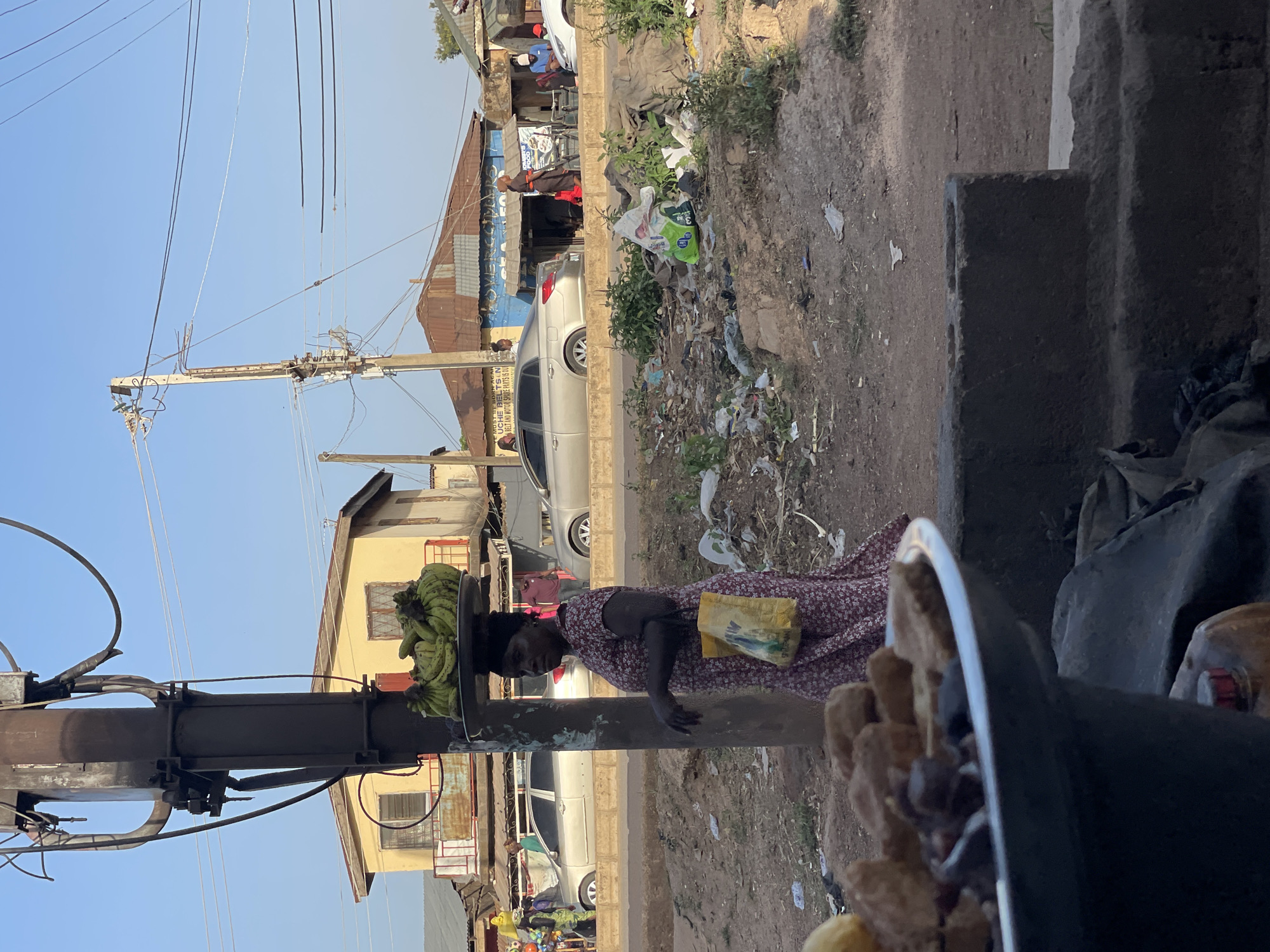 A medium-shot image, rotated vertically, depicts a woman in Bukuru, Nigeria, balancing a tray of green bananas on her head while walking along a street. The woman is wearing a red and white patterned dress and has a yellow bag slung over her shoulder. The street is littered with trash and debris. In the background, there are buildings, cars, and people. A black, metal object occupies the bottom of the frame. Overhead wires and utility poles are visible.