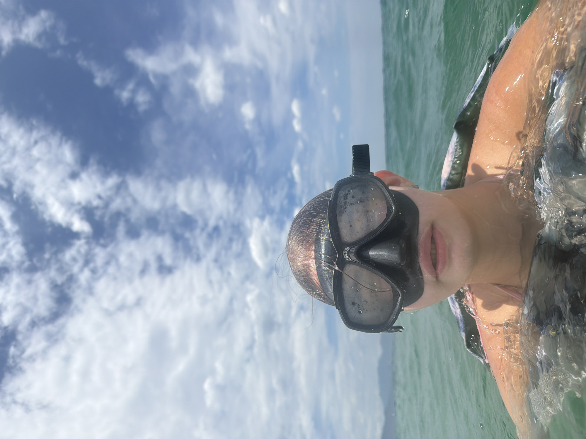A young person wearing a black Cressi diving mask and snorkel is swimming in the ocean. The person's head and shoulders are visible above the water, with the mask covering their eyes. The water is a clear turquoise color, and the sky is a bright blue with scattered white clouds. In the background, a distant shoreline with hills is visible. The scene appears to be a sunny day, likely in a tropical location. While the image does not contain explicit visual cues to pinpoint the exact location as Nha Trang, Vietnam, the general description of a coastal area with clear water, blue skies, and distant hills is consistent with the geography of many beaches in and around Nha Trang.