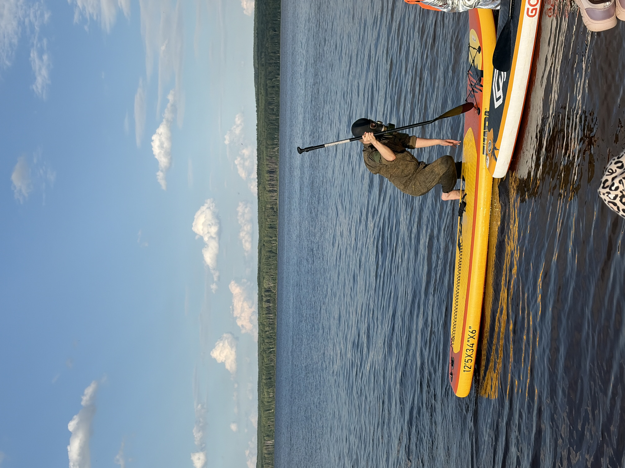 A person wearing a gas mask and a camouflage jumpsuit is on a yellow paddleboard in a lake. The person is holding a paddle and appears to be in the process of standing up. Another paddleboard, black and white, is next to the yellow one. The lake is calm with ripples on the surface, and a dense forest lines the distant shore under a blue sky with scattered clouds. The image is taken from a low angle, looking across the water. This scene is likely at the Kama River reservoir, near Chaikovsky, Russia, given the landscape.