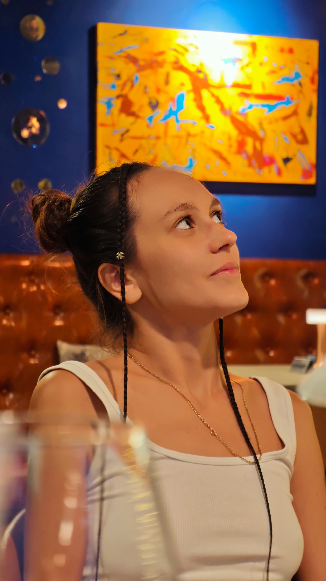 A young woman with dark hair styled in a top bun and two braids is looking upwards. She is wearing a white tank top and a delicate gold necklace. She has a clover-shaped charm attached to one of her braids near her ear. Behind her, a vibrant abstract painting with shades of orange, yellow, and blue is displayed on a dark blue wall. There are also several circular, reflective objects on the wall to the left of the painting. The background also features a textured, orange upholstered surface, possibly a booth or chair. In the foreground, out-of-focus elements suggest a table setting with glassware. The lighting is warm and dramatic, with a bright light source illuminating the painting from above. The image appears to be taken indoors, in what could be a restaurant or a stylish lounge. There is no identifiable information about the location within Hanoi, Vietnam.