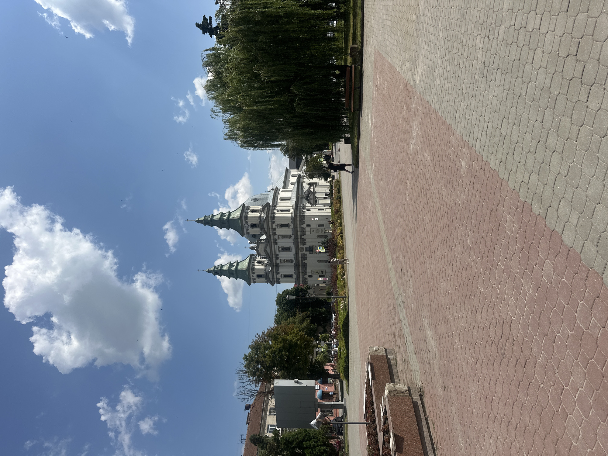 The image captures an outdoor plaza in Ternopil, Ukraine, dominated by a grand white church with two green-topped spires. A large, weeping willow tree stands prominently on the right side of the frame, its branches reaching downwards. The plaza's ground is paved with red and grey interlocking bricks, arranged in a hexagonal pattern. In the foreground, a concrete planter box with reddish granite accents is visible. Further back, a few people are present; one individual, dressed in dark clothing, is walking away from the viewer towards the right. Another person is seen in the distance near the church. The sky above is a clear blue with scattered white clouds. The overall scene depicts a sunny day in a public space adjacent to a significant religious building.