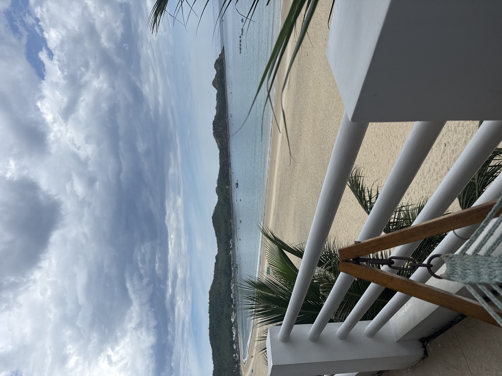 A wide shot of a beach viewed from a balcony. In the foreground, a white railing runs horizontally across the frame, with parts of a wooden-framed hammock with blue rope visible. Beyond the railing is a light-colored sandy beach. In the mid-ground, the ocean is a calm, light blue. Several small boats are scattered on the water. In the background, a range of green, tree-covered hills form the coastline. The sky is filled with dramatic, grey and white clouds.