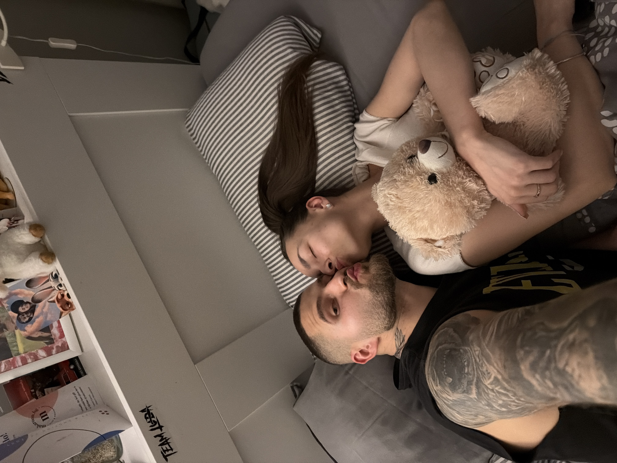 A couple is lying in bed, kissing. The woman is holding a teddy bear. The man has tattoos on his arm and neck. The location is likely indoors in a residential space. There is no indication of it being Sofia, Bulgaria.