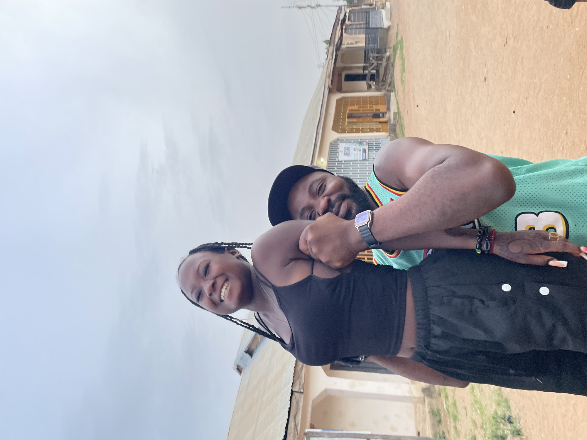 A dark-skinned woman with braids and wearing a black tank top and black pants is smiling and holding up her fists. A man with a beard and wearing a green jersey and a black baseball cap is standing behind her with his arms around her. They are standing outside in a city in Jos, Nigeria. The sky is overcast. In the background are buildings and dirt.
