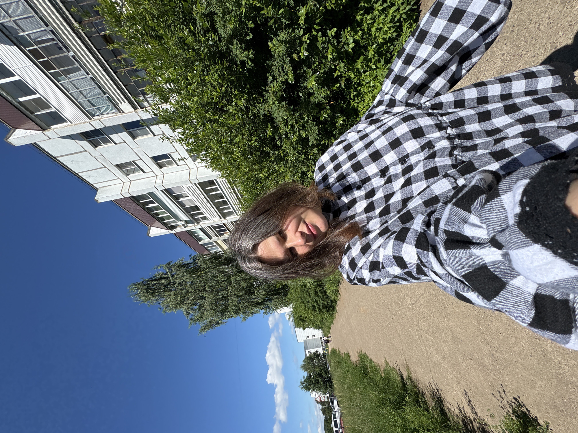 A woman with shoulder-length graying brown hair is captured in a selfie outdoors. She is wearing a black and white checkered shirt with puffed sleeves and a gathered waist. She is standing on a dirt path next to a grassy area. Behind her, a tall, slender tree stands to the left, and a multi-story apartment building with white siding and brown accents is visible to the right. The sky is a clear blue with a few scattered clouds. Further down the path, other buildings and a parked car can be seen. The overall scene depicts a sunny day in a residential area.