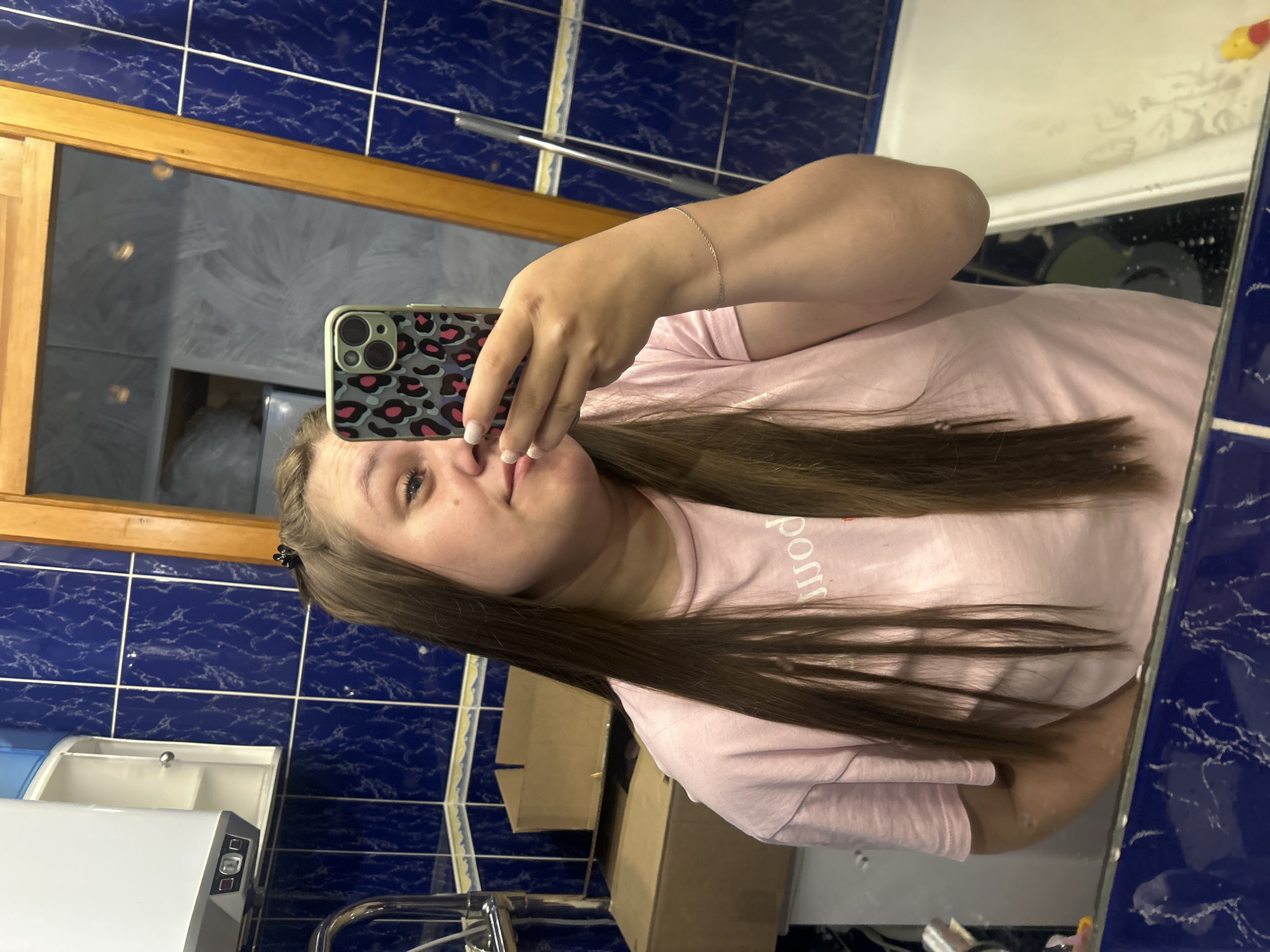 A woman is taking a selfie in a bathroom with blue tiled walls. She is wearing a pink t-shirt and has long brown hair. The bathroom has a mirror, a sink with a white cabinet, and a towel rack. The reflection in the mirror shows a window or doorway and some other objects in the bathroom. The exact location in Chaikovsky, Russia is not discernible from the image.