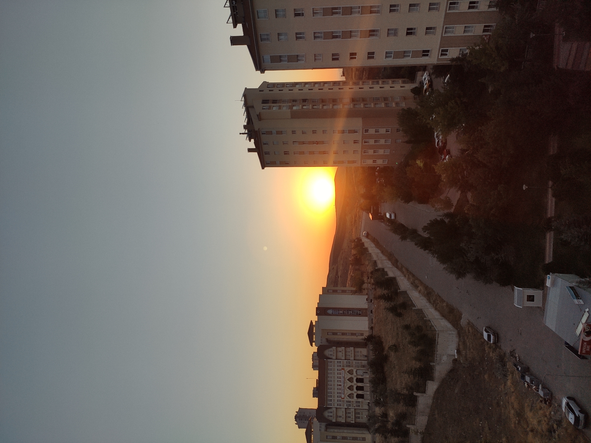 The photograph captures a sunset scene in Etimesgut, Turkey, with an expansive sky transitioning from a pale blue to a warm orange glow near the horizon. Two modern, tall residential buildings dominate the right side of the frame, characterized by their cream-colored facades and numerous windows. To the left, a large, multi-story institutional building, possibly a school or government facility, stands with a more traditional architectural style, featuring multiple rows of windows and an arched entrance. A paved road runs through the middle ground, flanked by sparse, dry vegetation and a low concrete wall. Several cars are parked along the road and in a lot to the left of the institutional building. The overall environment suggests a suburban or semi-urban setting within Etimesgut. The lighting indicates it is late afternoon or early evening, with the sun low in the sky.