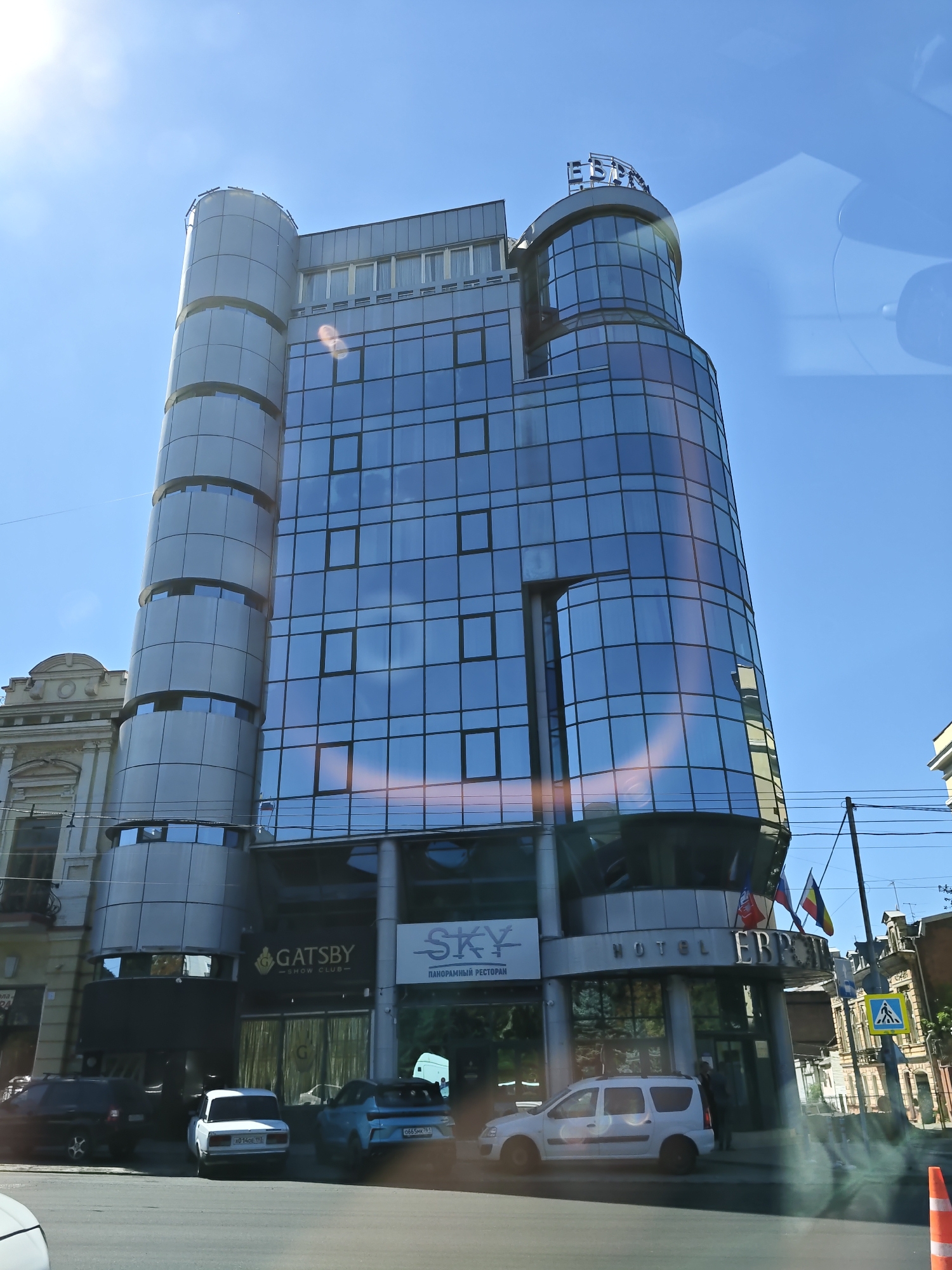 The image shows a modern, multi-story building with a glass facade in Rostov-on-Don, Russia. The building is labeled "HOTEL EBRI" at its entrance. Below the hotel entrance, there are signs for "GATSBY SHOW CLUB" and "SKY PANORAMIC RESTAURANT". Several cars are parked on the street in front of the building, including a black sedan, a white sedan, and a light blue sedan. A white minivan is also parked to the right. The sky is clear and blue with some sun glare. The scene appears to be captured from a vehicle.