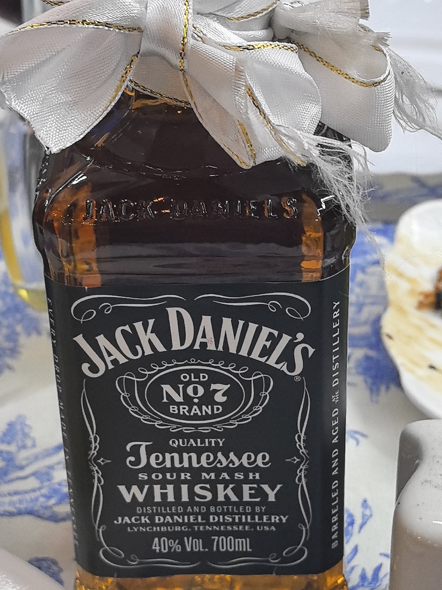 The image shows a close-up of a small bottle of Jack Daniel's Tennessee Whiskey. The bottle is dark amber and has a white bow with gold trim tied around its neck. The label is predominantly black with white lettering. The text on the label reads: "JACK DANIEL'S", "OLD NO. 7 BRAND", "QUALITY TENNESSEE SOUR MASH WHISKEY", "DISTILLED AND BOTTLED BY JACK DANIEL DISTILLERY LYNCHBURG, TENNESSEE, USA", and "40% VOL. 700mL". Vertical text along the side of the label reads: "BARRELED AND AGED AT THE DISTILLERY". The bottle is placed on a surface that appears to be a table covered with a blue and white patterned cloth. In the background, a blurred image of a white dessert, possibly a cake, can be seen. The scene is lit with bright light, creating reflections on the glass bottle. There is no indication of the city of Taldykorgan, Kazakhstan, in the image.