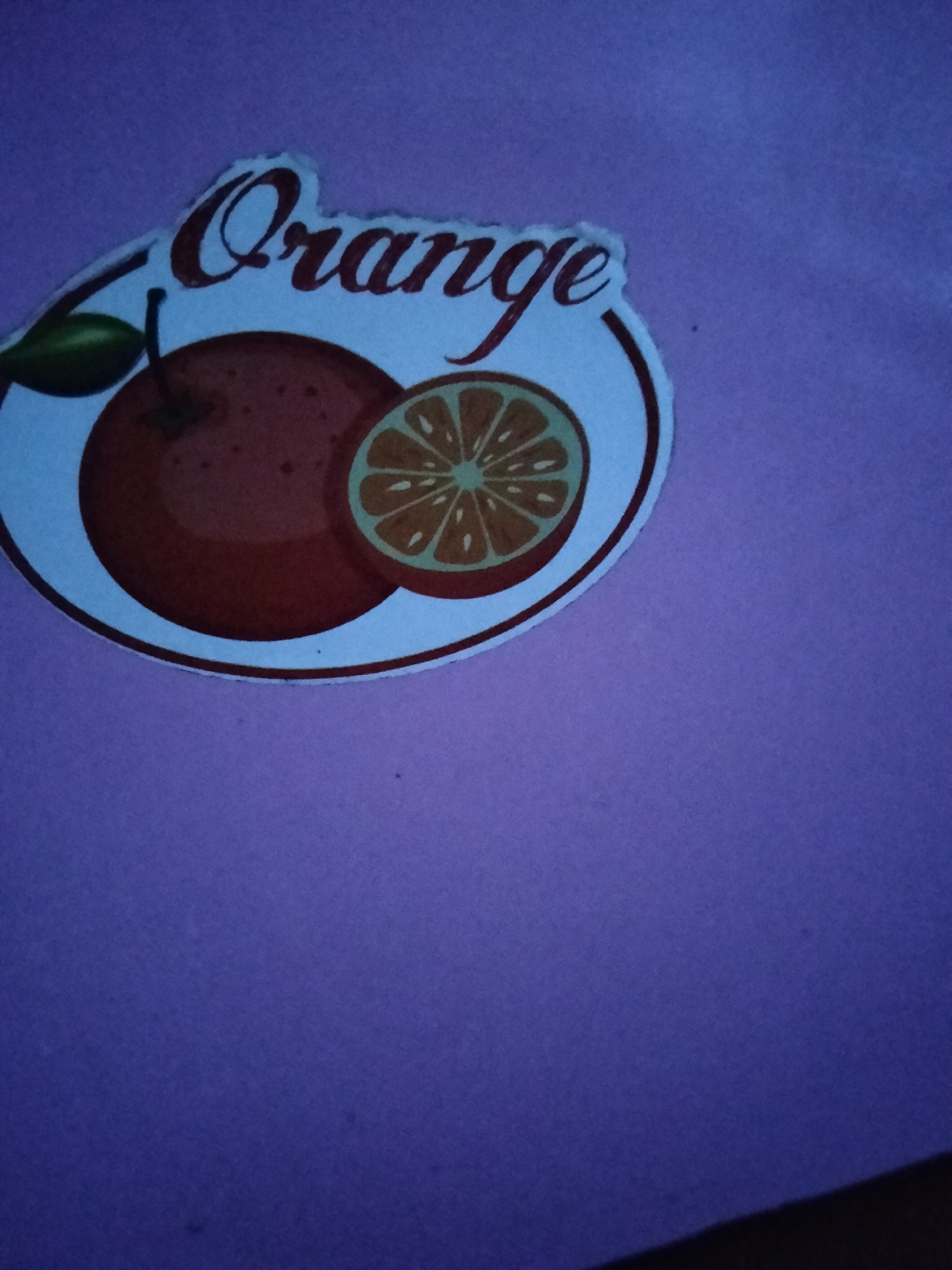 The image shows a sticker featuring an orange fruit and the word "Orange" in stylized text. The sticker is placed on a purple surface, possibly paper or fabric. The sticker itself is circular with a white border. It depicts a whole orange on the left and a cross-section of an orange on the right. The text "Orange" is written in a cursive font above the fruits. The purple surface appears to be a flat background. The lighting in the image is dim, casting some shadows and highlighting the texture of the purple surface. The scene is static and there are no people or active events depicted. The location in Kogon City, Uzbekistan, is not evident from the visual information provided in the image.