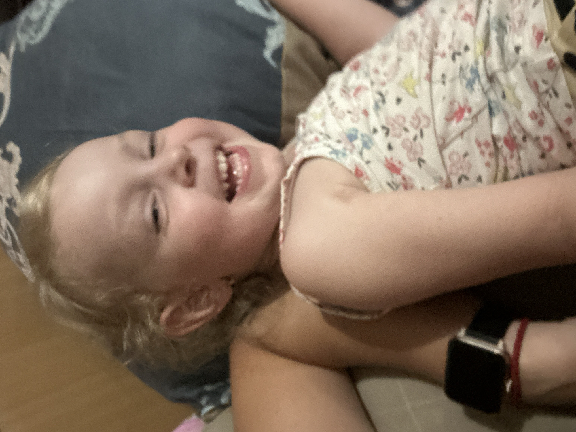 A close-up, angled shot shows a young child lying on their back, looking up and smiling broadly. The child has blonde hair and is wearing a white top with a floral pattern of pink, blue, and yellow flowers. Their mouth is open, revealing teeth. The child's head is resting on a dark blue patterned fabric, possibly a pillow or blanket. Another person's arm, wearing a black smartwatch with a red band, is visible in the lower right corner, indicating they are likely holding or interacting with the child. The background appears to be a wooden floor. The lighting suggests it is daytime, and the setting appears to be indoors. The child's expression conveys happiness and amusement.