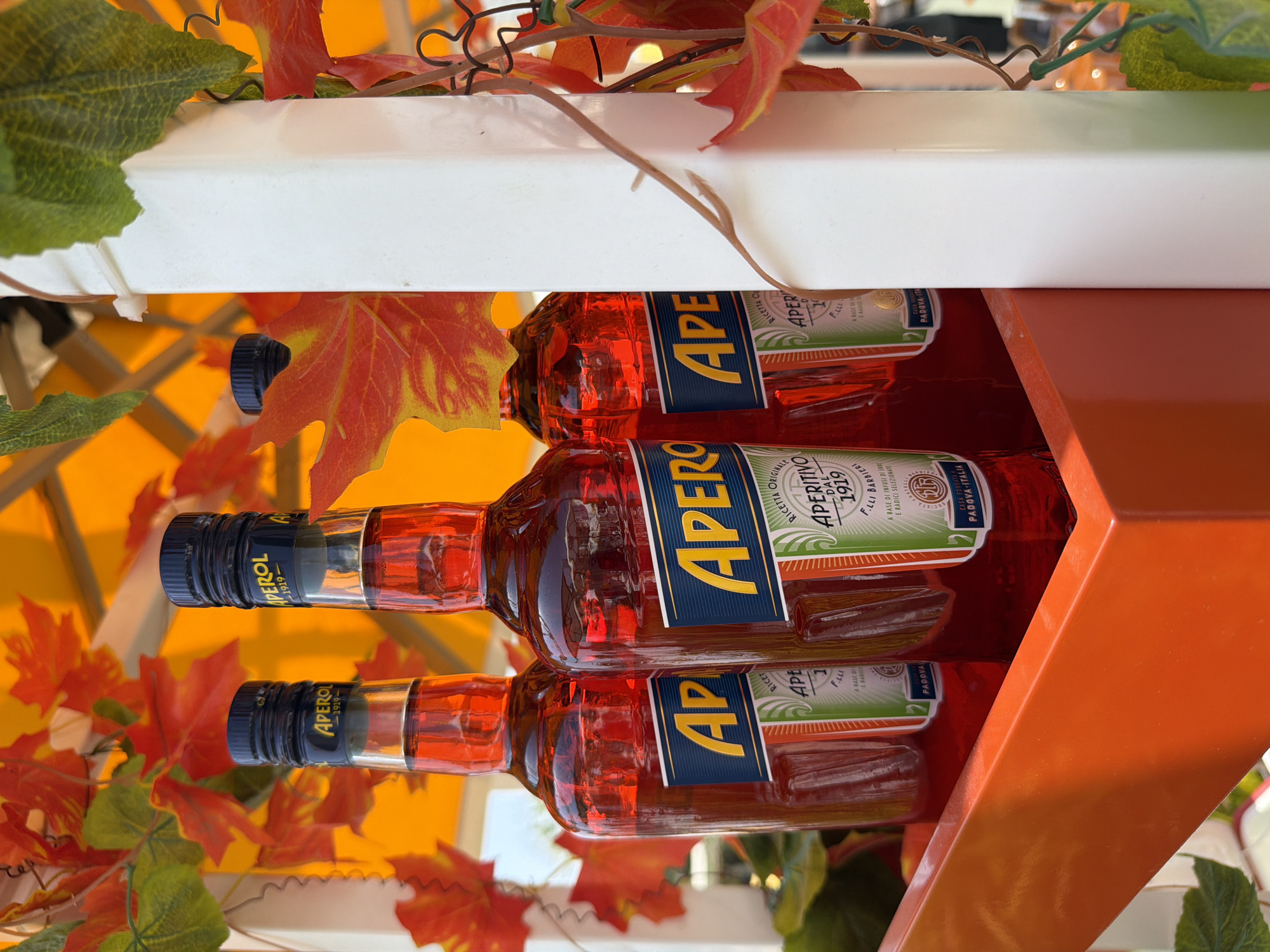 Three bottles of Aperol liqueur are displayed in a bright orange holder, with artificial autumn leaves and vines adorning the white structure behind them. The bottles are partially obscured by the leaves, and the scene is lit by an orange overhead awning. The background suggests an outdoor setting, possibly a bar or restaurant, though no specific location in Dubai is evident.